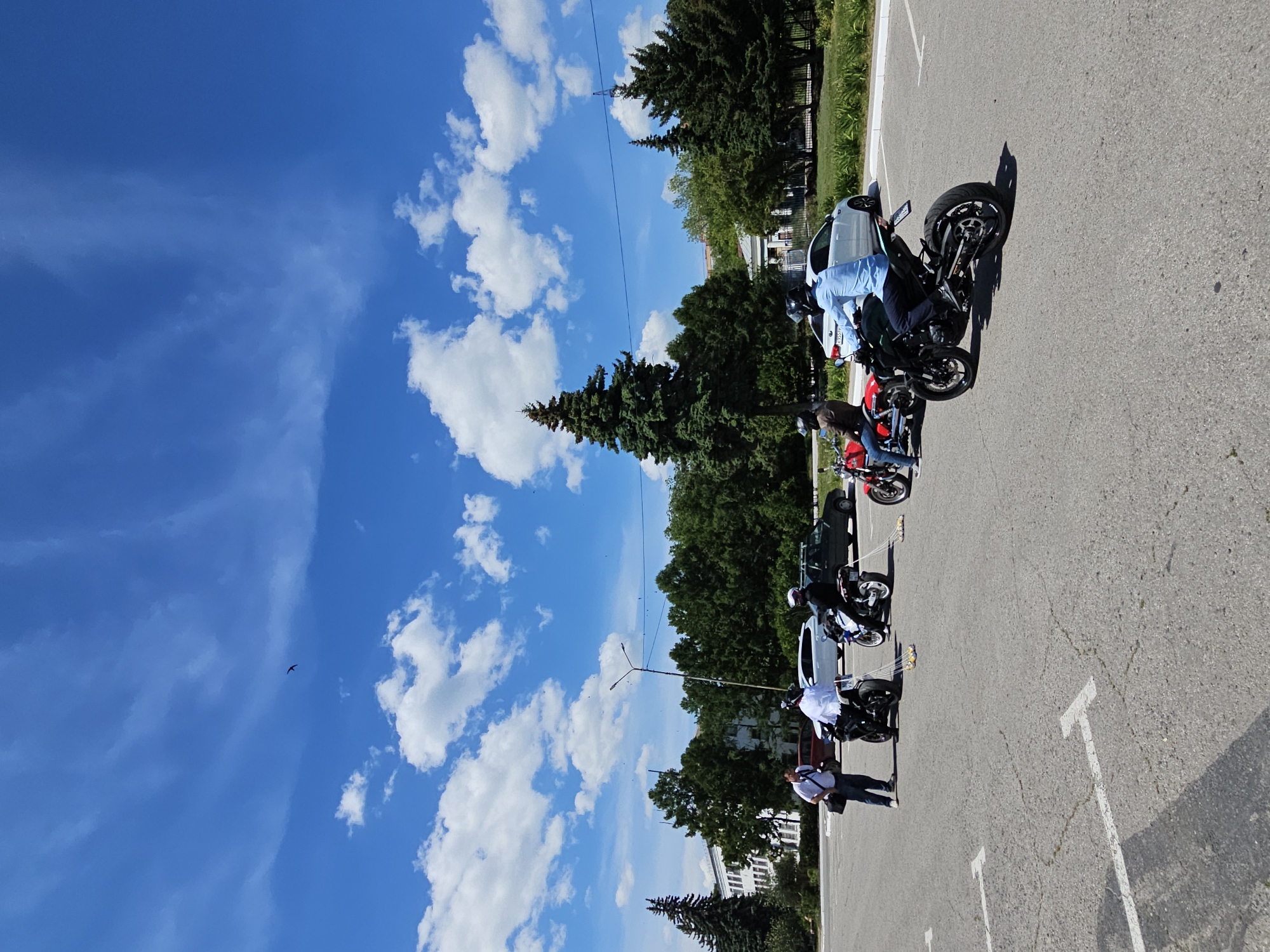 The image, taken in Ulyanovsk, Russia, depicts a sunny day with a bright blue sky dotted with white clouds. Several motorcycles are parked in a line on an asphalt lot, marked with white parking lines. A few people are visible, some near the motorcycles. One person in a blue shirt and dark pants is seated on a motorcycle, while another person in a white shirt and jeans stands nearby. Further down the line, another individual in a white shirt and dark pants is walking away from a motorcycle. The scene appears to be a casual gathering or a break during a motorcycle ride, possibly in a public parking area with trees and buildings in the background. The overall mood seems relaxed and pleasant, fitting for a clear, warm day. No visible text is present in the image.