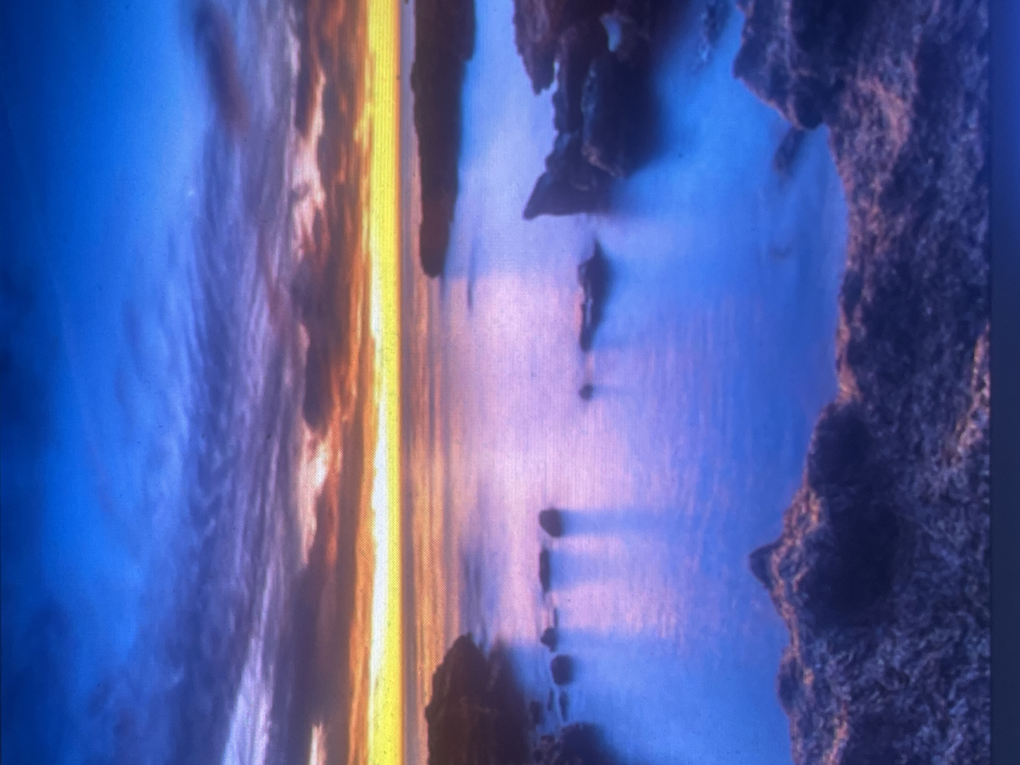 This image captures a vibrant coastal sunset or sunrise, depicted as being in the city of Zelenyi Hai, Ukraine. The scene features a body of calm water surrounded by dark, rugged rock formations that define the shoreline and appear as scattered islets within the water. The sky transitions from a deep, saturated blue overhead to lighter blue, purple, and then intense orange and yellow at the horizon, where the sun's glow is brightest. Stratus clouds, varying in density and color from dark gray-purple to orange and red, stretch across the middle sky. The placid water vividly reflects the sky's colors, appearing as gradients of purple, pink, and blue, with elongated shadows cast by the rocks on its surface. There are no individuals, vehicles, or visible human structures in the frame, presenting a serene, natural landscape. The entire image has a visible pixel grid, suggesting it is a photograph of a digital display.