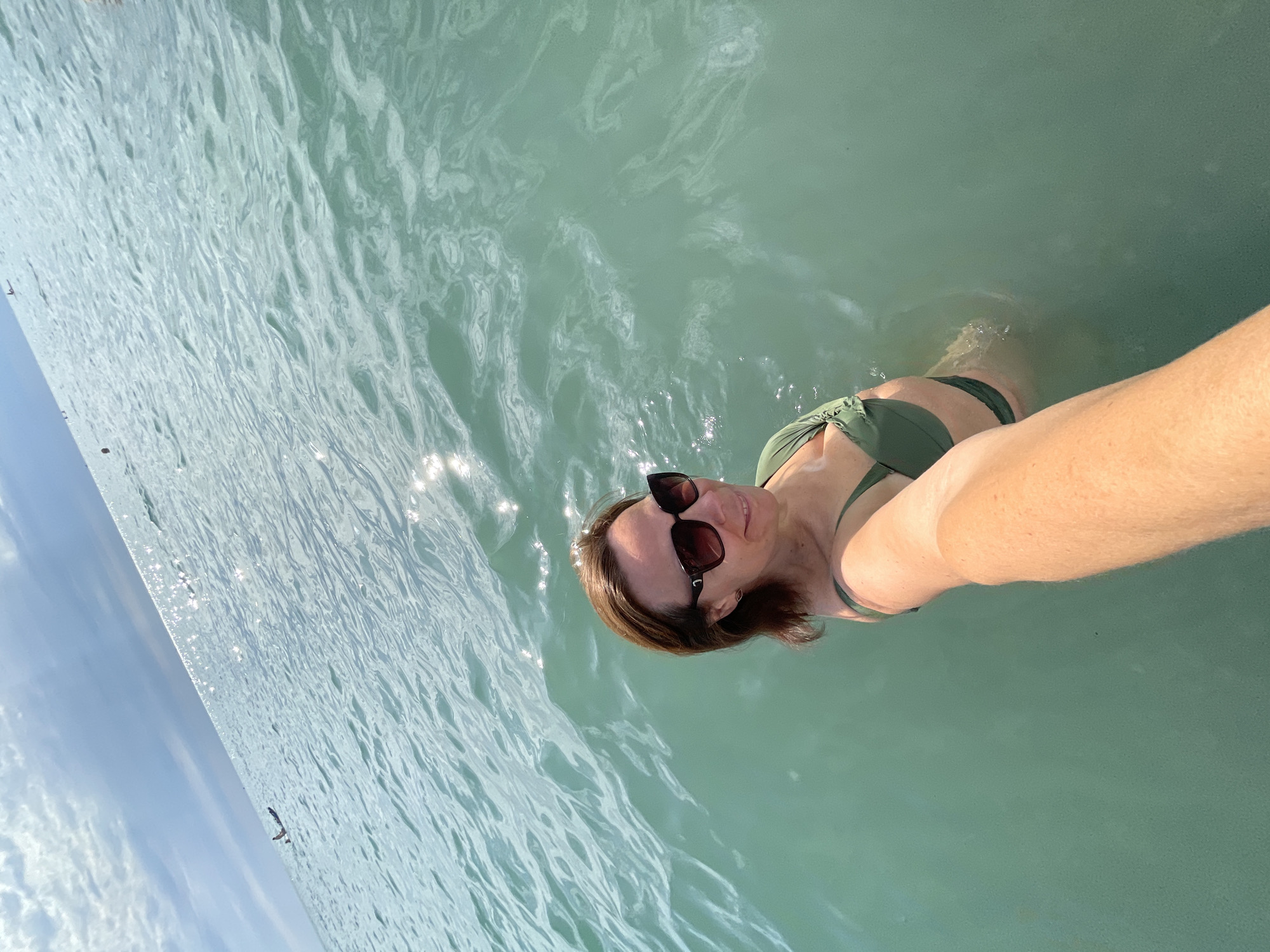 A woman in a green bikini and sunglasses takes a selfie while standing in the sea. The water is clear and blue-green, with sunlight reflecting off the surface. The sky is partly cloudy. In the distance, there are a few small boats and buoys. The image appears to have been taken on a sunny day.