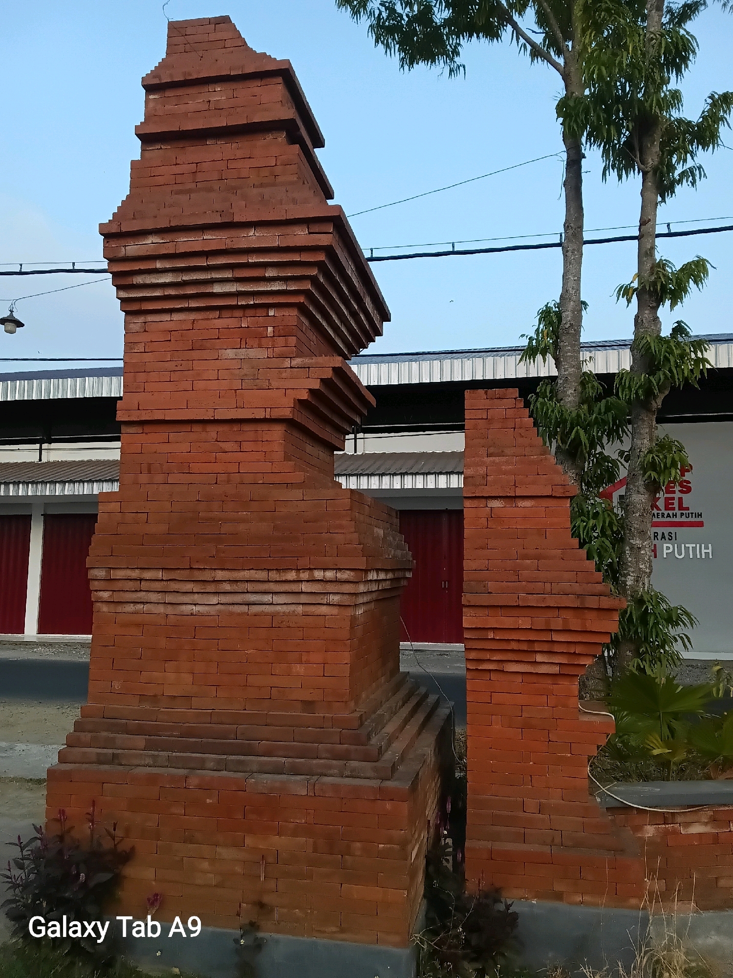The image shows a brick structure in Bendorejo, Indonesia, under a clear blue sky. The structure is made of red bricks and has a tiered design, resembling a gate or an entrance. It is partially broken on the right side, revealing the interior construction. Behind the brick structure, there are two trees with green leaves and a building with red doors and a white facade. A sign with text in Bahasa Indonesia is visible on the building's facade. In the foreground, there are some small plants and the text "Galaxy Tab A9" is present at the bottom left corner. The overall scene suggests an outdoor setting, possibly a public area or a historical site.