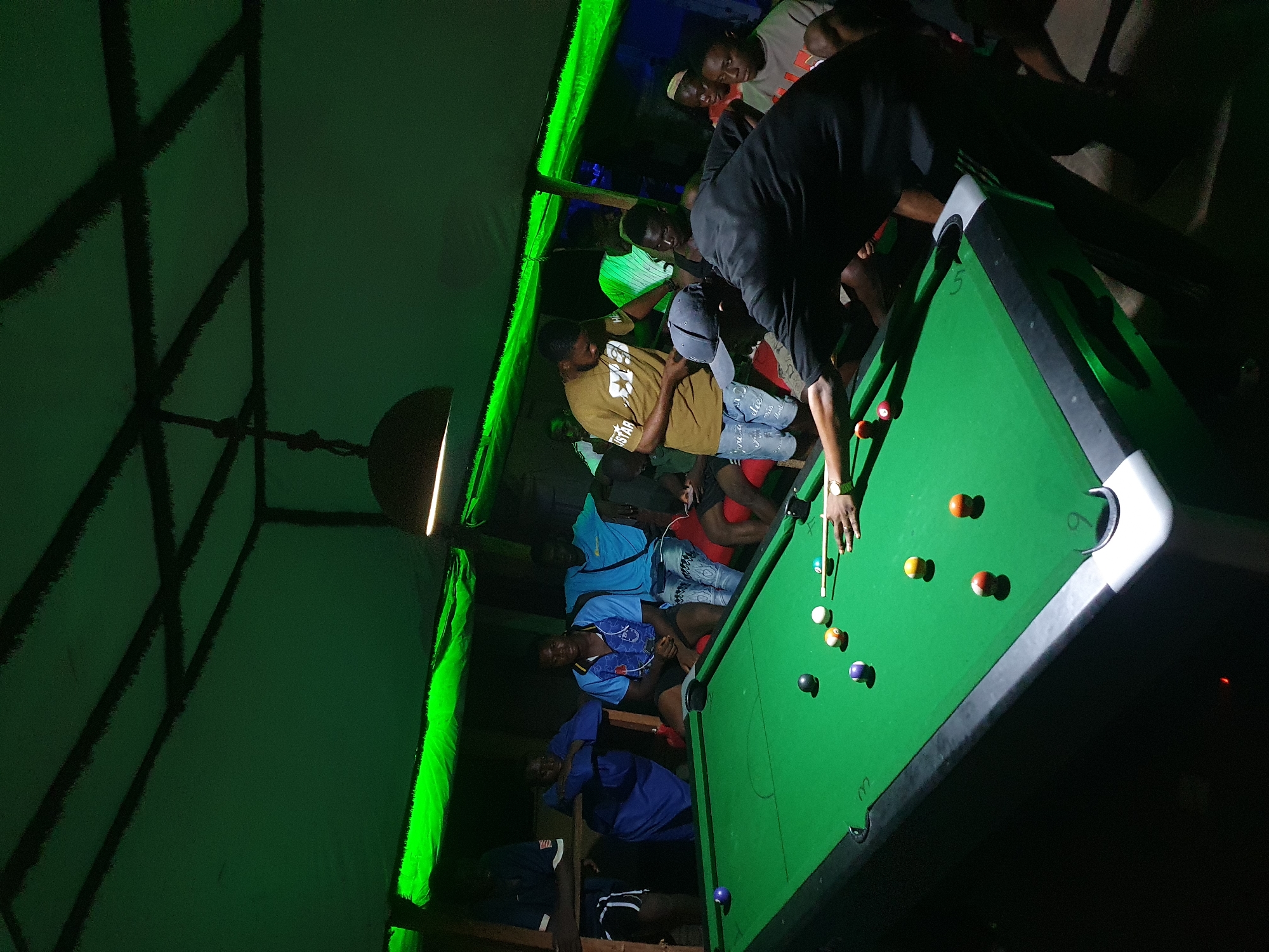 The image captures an indoor scene, likely at night, in Lokoja, Nigeria, illuminated primarily by green ambient lighting. The main subject is a group of young men gathered around a pool table.
One man in a black shirt is actively playing pool, leaning over the green felt table, holding a cue stick, and aiming at the billiard balls. Several colored balls are scattered on the table, including solids and stripes, with the cue ball positioned for a shot. Another person's hand is also visible near the cue stick.
Around the pool table and along the wall, other men are seated or standing, observing the game and socializing. They are dressed in casual attire, including t-shirts and jeans. One man wears a mustard-colored t-shirt with "STAR" visible on it, while another wears a blue shirt with a pattern. Some are wearing caps. The seating appears to be wooden benches or platforms.
Overhead, a lamp fixture hangs, casting a localized light. The ceiling or upper wall features a dark, crisscrossing pattern, possibly from structural beams or decorative elements, contrasted against the green-lit surface. The overall atmosphere appears casual and social, focusing on the game of pool as a central activity. The image is taken from an elevated perspective, looking down at the scene, and is rotated counter-clockwise.