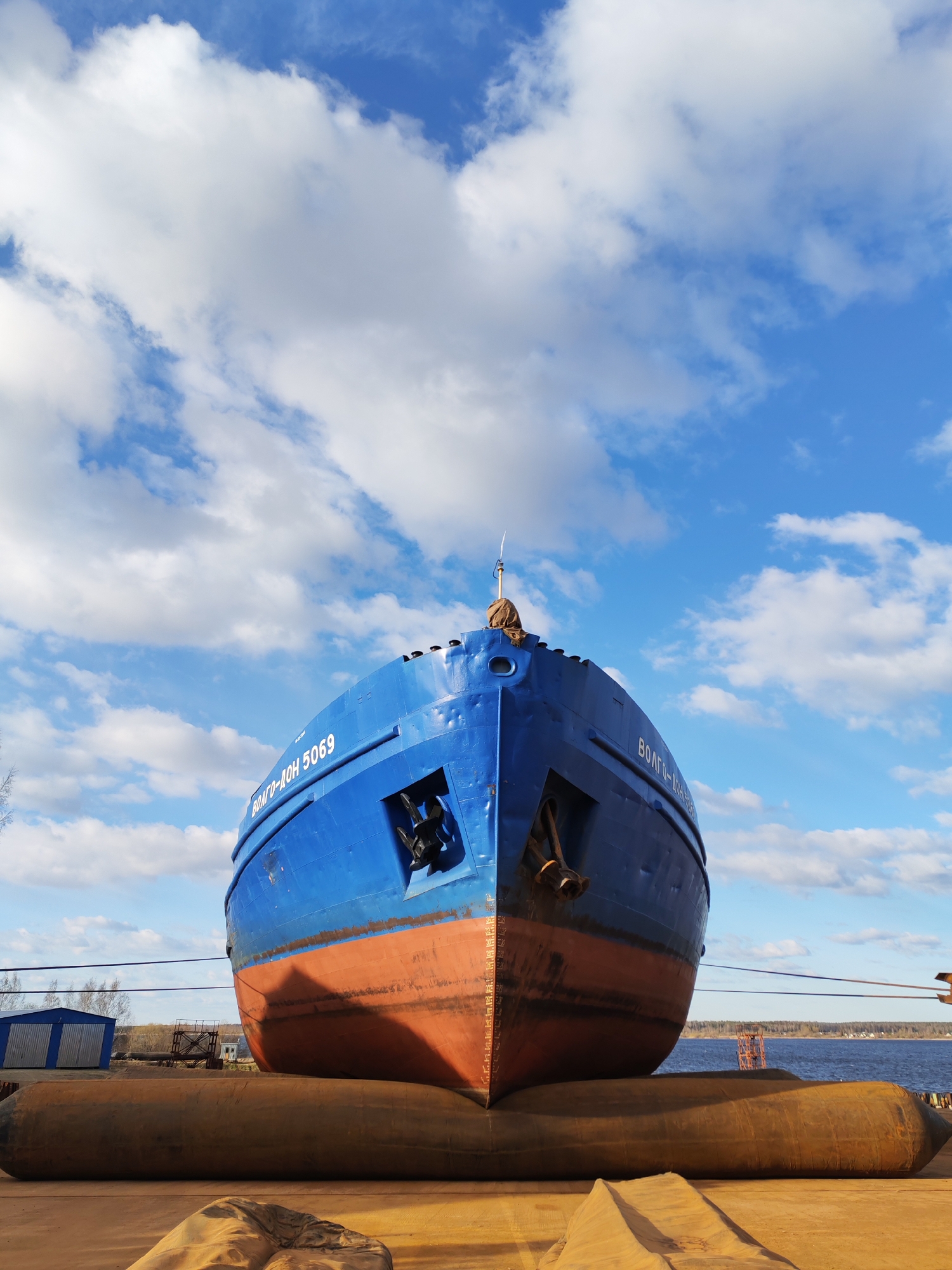 A large ship, identified by the Cyrillic text "ВОЛГО-ДОН 5069" (VOLGO-DON 5069) on its bow, is depicted out of the water in what appears to be a shipyard or dry dock in Bol'shie Mkhi, Russia. The ship's prominent bow section is painted a vibrant blue above the waterline and a rust-red below, showing visible signs of weathering and rivets. Two large anchors are partially visible in their housing on the ship's bow.
The vessel rests on several large, cylindrical, yellow-brown inflatable airbags or rollers, suggesting it is either being moved across land, prepared for a slipway launch, or positioned for maintenance. Support lines or cables extend from the ship's hull to the foreground.
The setting is outdoors on a bright, clear day, with a vivid blue sky adorned with fluffy white cumulus clouds dominating the upper portion of the frame. In the background, a body of water, likely a river or canal, stretches horizontally, bordered by a distant, tree-lined bank. To the left, glimpses of industrial sheds or buildings are visible. The time of day appears to be late morning or afternoon, given the bright, direct sunlight. No individuals are visible in the image. The scene captures an active industrial environment focused on maritime operations.