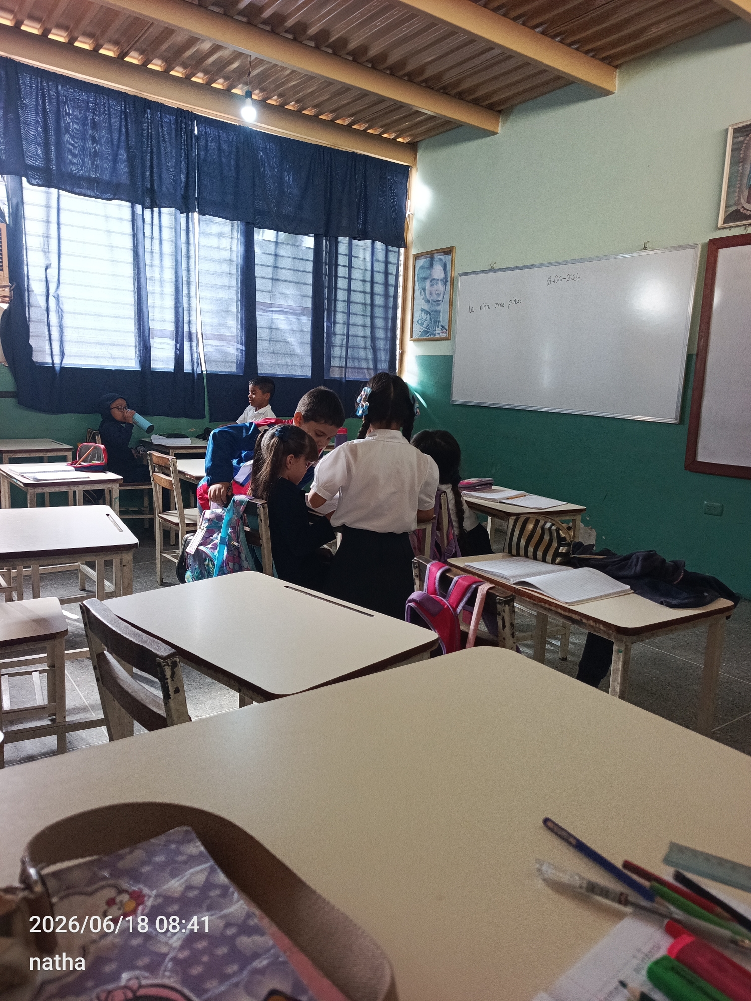 The image depicts an indoor scene, likely a classroom in San Sebastián, Venezuela, given the timestamp. Several students are visible at their desks. In the foreground, a table with writing utensils, including pens and a ruler, is in focus. The middle ground shows students engaged in classroom activities. One student, wearing a white shirt, appears to be interacting with another student seated at a desk. Other students are also at desks, some with backpacks and notebooks. In the background, a whiteboard is visible with writing on it, and a portrait is hanging on the wall. The room has windows with dark blue curtains and exposed ceiling beams. The lighting suggests it is daytime.
