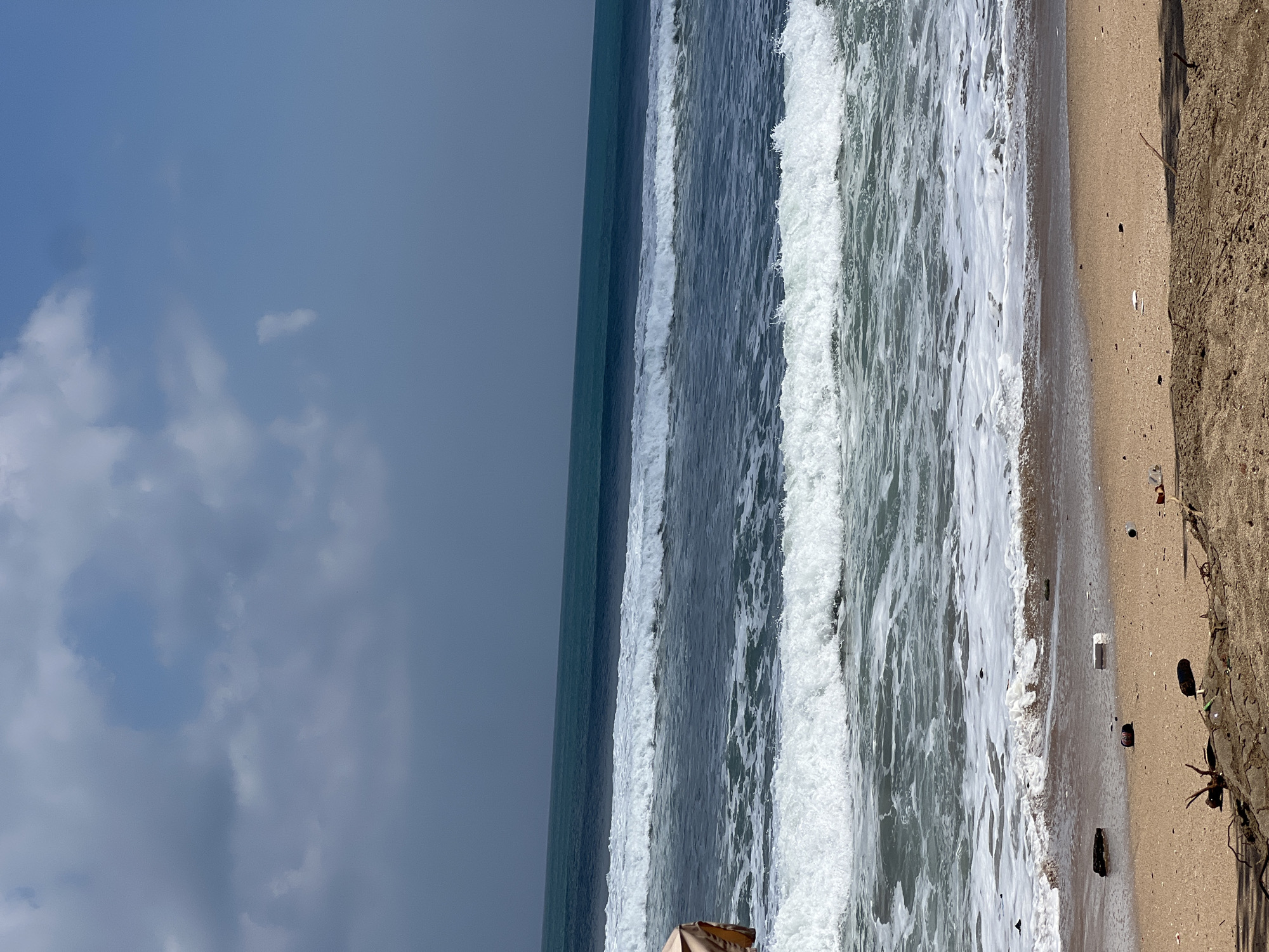 The image captures a wide view of a beach in Kuta, Indonesia, with the ocean dominating the frame. Several waves are rolling in, creating white foam as they crash onto the shore. The water transitions from a deep blue in the distance to a lighter shade closer to the beach, with multiple layers of waves visible. The sky above is a mix of blue and grey clouds. The sandy beach in the foreground is littered with various pieces of debris, including plastic bottles, wrappers, and driftwood. A small portion of a tan-colored umbrella is visible on the far left of the image, and a person is partially visible underneath it. The overall scene depicts a natural coastal environment impacted by human activity, evidenced by the scattered trash on the sand.