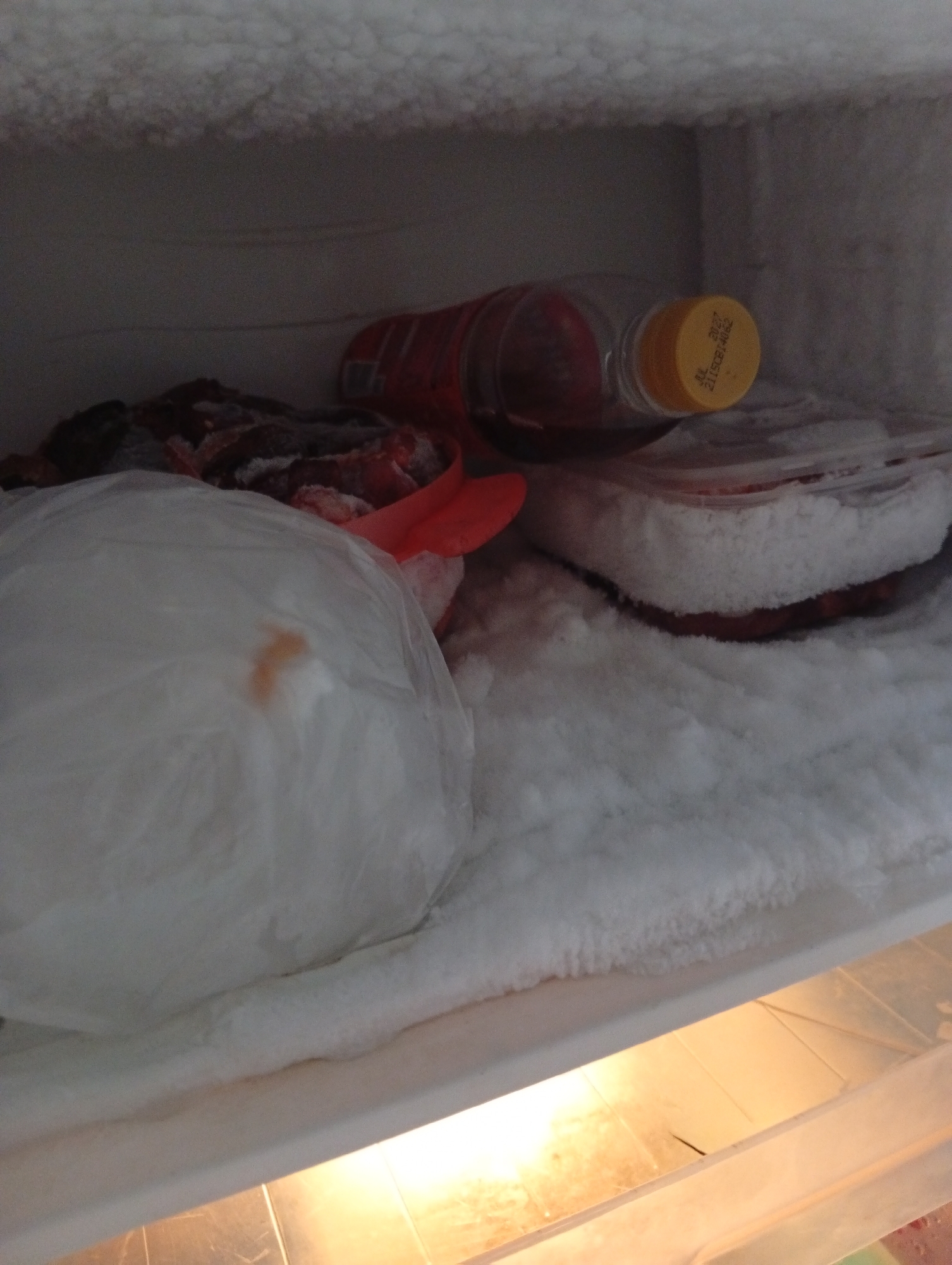The image depicts the interior of a frost-covered refrigerator compartment, likely in a residential kitchen setting within Kualu, Indonesia. The foreground features a large, white, translucent plastic bag containing some frozen food items, with a small orange stain visible on its surface. To the right and behind this bag, a red plastic utensil is partially obscured. Further back, a dark red liquid is contained within a clear plastic bottle with a yellow cap. The bottle is lying on its side, and a portion of its label is visible, displaying some text and numbers. The background is dominated by the frosty walls of the refrigerator compartment. The overall environment suggests items have been stored for an extended period, indicated by the accumulation of frost. There are no individuals present in the image, and the focus is solely on the frozen contents and the interior of the refrigerator.