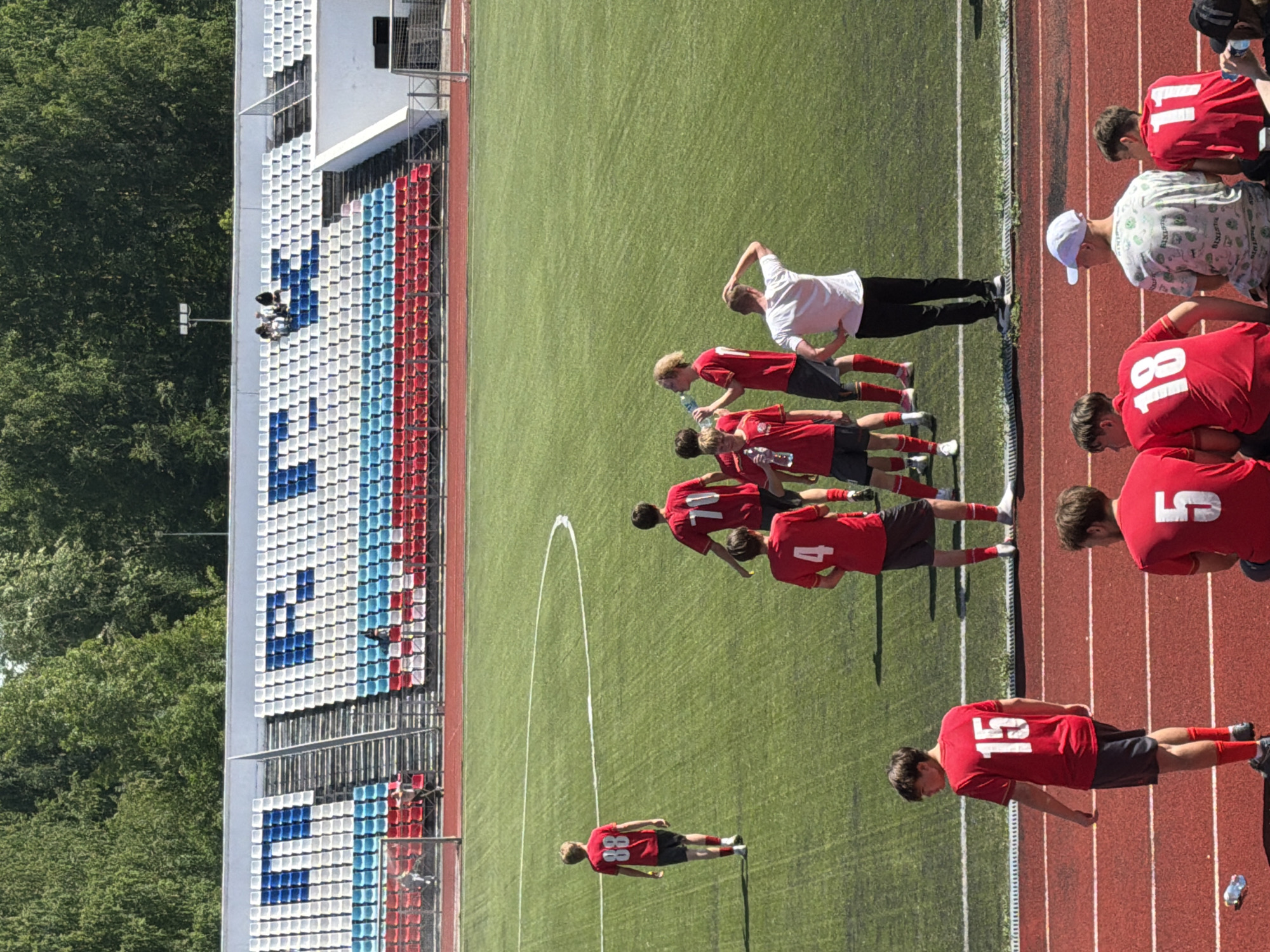 The image captures an outdoor daytime scene at Energiya Stadium in Chaikovsky, Russia. The stadium features a green artificial turf soccer field surrounded by a reddish-brown running track. The stadium grandstands in the background display seating arranged in white, blue, and red colors, spelling out "STP" on the left side (partially obscured by a goal) and "ENERGIYA" on the right side using blue seats against a white background, with lower sections of light blue and red seats. Dense green trees are visible behind the stadium structure under a bright, clear sky.
On the field, approximately seven young males, presumably soccer players, are present. Most wear red short-sleeved jerseys with white numbers and dark shorts. One player (number 88) walks away from the center toward the left. A group of players gathers near the center-right; one (number 70) is seen from behind, another (number 4) is partially visible, and one with blonde, curly hair drinks from a plastic water bottle. An adult male in a white t-shirt and dark pants stands observing this group, with one hand on his head and the other on his hip.
In the immediate foreground, on the running track, are five individuals. One young male (number 15) stands facing away, wearing a red jersey and dark shorts. Three other young males are seated: one (number 5) in a red jersey, one (number 18) in a red jersey, and one between them wearing a white baseball cap and a light-colored, patterned t-shirt. A fourth seated individual (number 11) in a red jersey is partially visible on the right. A clear plastic water bottle lies discarded on the track near player 15.
A few spectators are sparsely distributed in the upper sections of the grandstands, with approximately three visible in the "ENERGIYA" section and one in the "STP" section. Bright sunlight casts distinct shadows across the field and track.