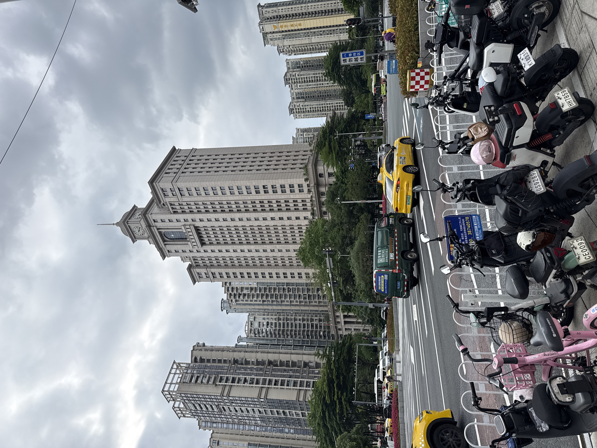 This is a street-level, eye-level, medium shot of a city street in Tianhe District, China. The scene is dominated by a large, ornate building with a clock tower, likely a government or official building, and several modern, high-rise residential buildings in the background. The sky is overcast with gray clouds.
In the foreground, several electric scooters and motorcycles are parked along a white metal barrier. One pink electric scooter stands out, equipped with a basket. Further back, a street with multiple lanes of traffic is visible. Several yellow taxis and a green delivery van are on the road, along with other vehicles. Pedestrians are present but not prominently featured. Traffic lights and street signs are also visible, indicating an urban environment. The overall impression is a bustling city scene on a cloudy day.