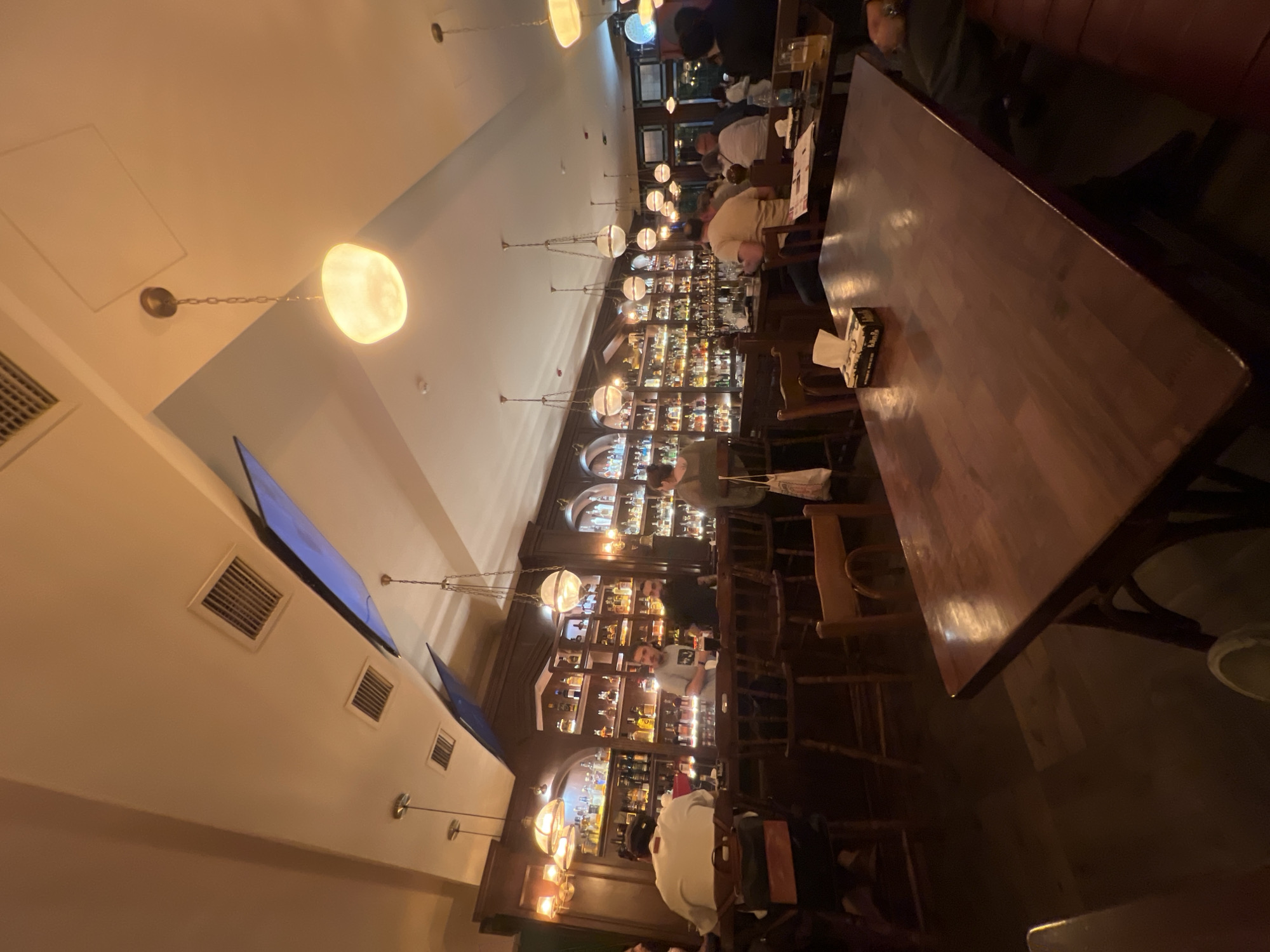 The image depicts the interior of a bar or restaurant in Yerevan, Armenia. The establishment features a long, polished wooden bar stocked with numerous bottles of alcohol, illuminated by warm lighting. Behind the bar, several staff members are visible, along with patrons seated on stools. The ceiling is high, with exposed ventilation systems and several decorative globe-shaped light fixtures hanging from chains. The overall ambiance is dimly lit and cozy, with wooden tables and chairs in the foreground and middle ground, occupied by customers. The flooring appears to be dark wood.