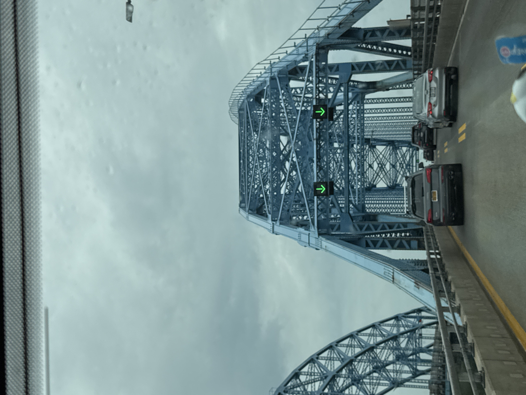 The image shows a view from inside a vehicle traveling on a bridge, likely during the day under overcast skies. The bridge is a large steel truss structure, painted blue, and appears to be quite old. Traffic is visible on the bridge, with several cars driving ahead. Green arrow traffic lights are visible, indicating the direction of traffic flow. A portion of the vehicle's windshield wiper or frame is visible on the left side of the image. The sky is a uniform gray, suggesting cloudy weather, possibly with rain or fog. The road surface is asphalt with yellow lane markings. There is no visible text in the image. The scene evokes a sense of travel and transit, with the imposing bridge structure and the steady flow of vehicles. The location is identified as the Town of Tonawanda, United States of America.