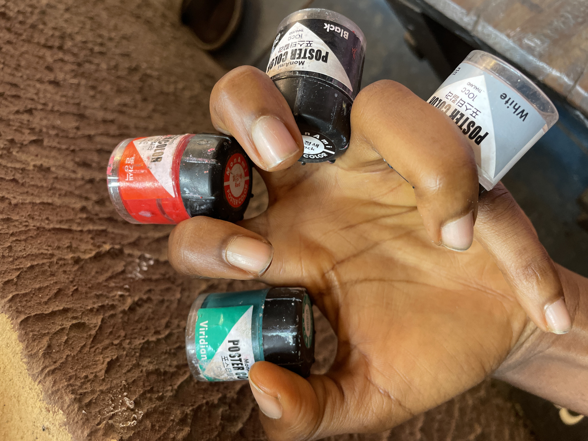 An arm holds four small paint jars labeled MonAmi Poster Color. The colors are Viridian, Red, Black, and White. The paint jars appear to be made in Thailand. The background is blurry and textured. The location in the city of Nkpor, Nigeria, is not discernible from the visual data.