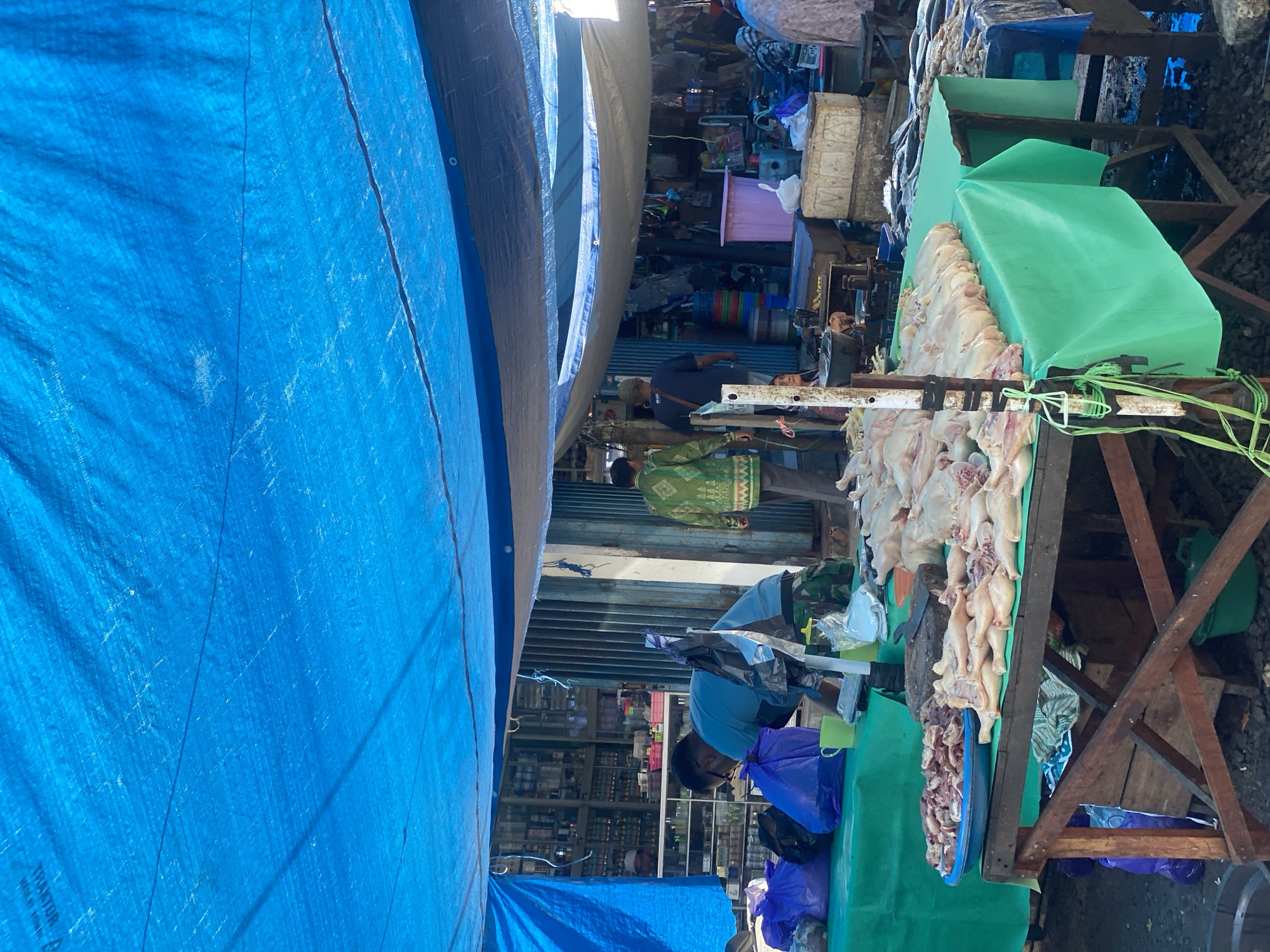 A busy outdoor market scene is captured under a large blue tarpaulin. The market appears to be a food market, with several tables displaying fresh produce and raw meats. In the foreground, a table covered with a green cloth showcases a large quantity of raw chickens laid out neatly, along with a blue bowl filled with cut-up pieces of meat. Another table to the left displays more meat items.
Several individuals are visible in the scene. A man wearing a blue t-shirt is leaning over a table, possibly preparing or arranging items. Another man in a green patterned shirt and dark pants walks away from the camera. Further in the background, a man in a dark shirt is seen walking.
Behind the market stalls, shelves stocked with various items, likely spices or other market goods, are visible through an open doorway. The overall environment suggests a lively and active marketplace. The location is identified as Desa Tunggul Irang, Indonesia.