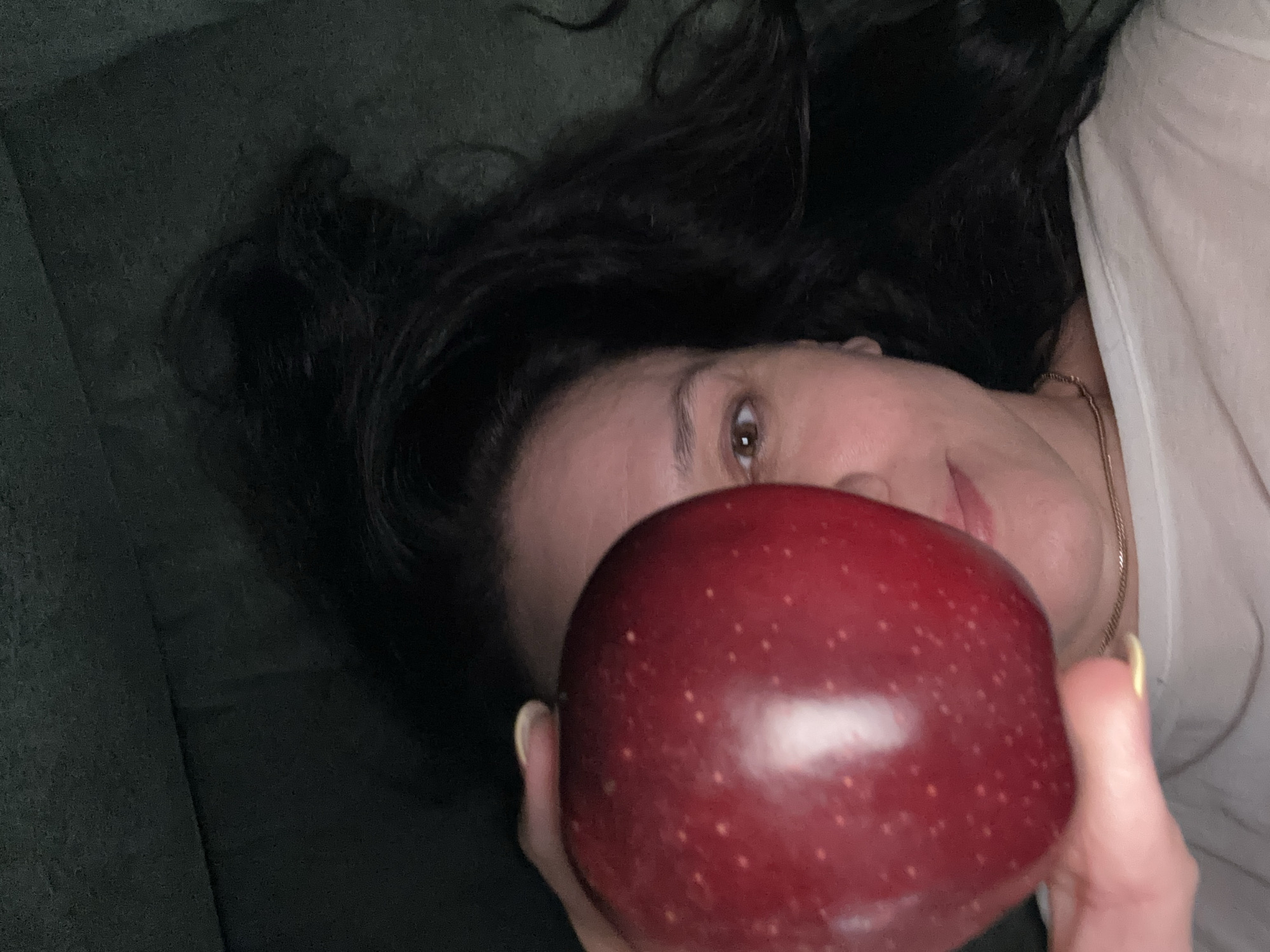 A person with dark hair is lying down, holding a red apple in front of their face. The person appears to be wearing a white shirt and a gold chain. The background is a dark green or gray color. It is not possible to determine the location.