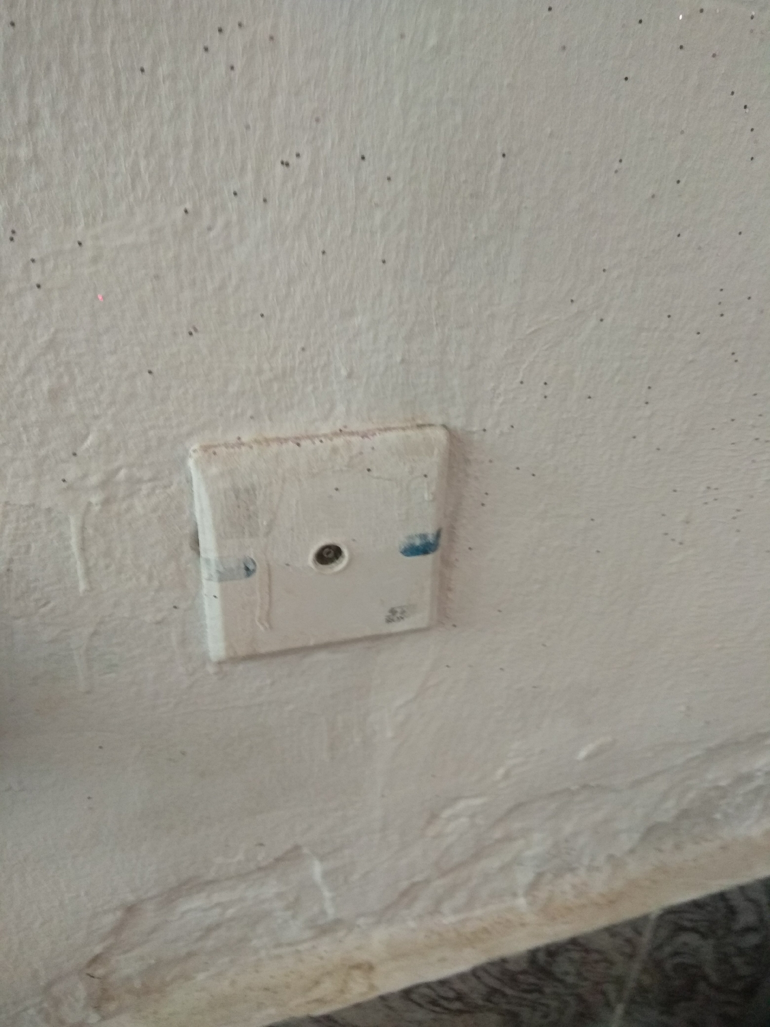 The image shows a close-up of a white wall in Jalingo, Nigeria. The wall has a textured surface with numerous small, dark specks, possibly dust or insects, scattered across it. There are also some faint white drips or streaks running down the wall.
Affixed to the wall is a white, square electrical or data plate. The plate appears to be somewhat aged and has some blue marks on it, possibly from paint or marker. Centered on the plate is a circular socket, which appears to be a coaxial connection for cable television or an antenna. The plate is slightly recessed into the wall, and the edges of the plate are visible.
In the lower part of the image, a sliver of a patterned floor is visible, with dark and light grey wavy lines. The lighting suggests it is daytime, but there are no clear indications of time or weather conditions. The overall impression is of an interior wall, likely in a residential or commercial building, with an older, somewhat worn aesthetic. There is no visible text in the image.