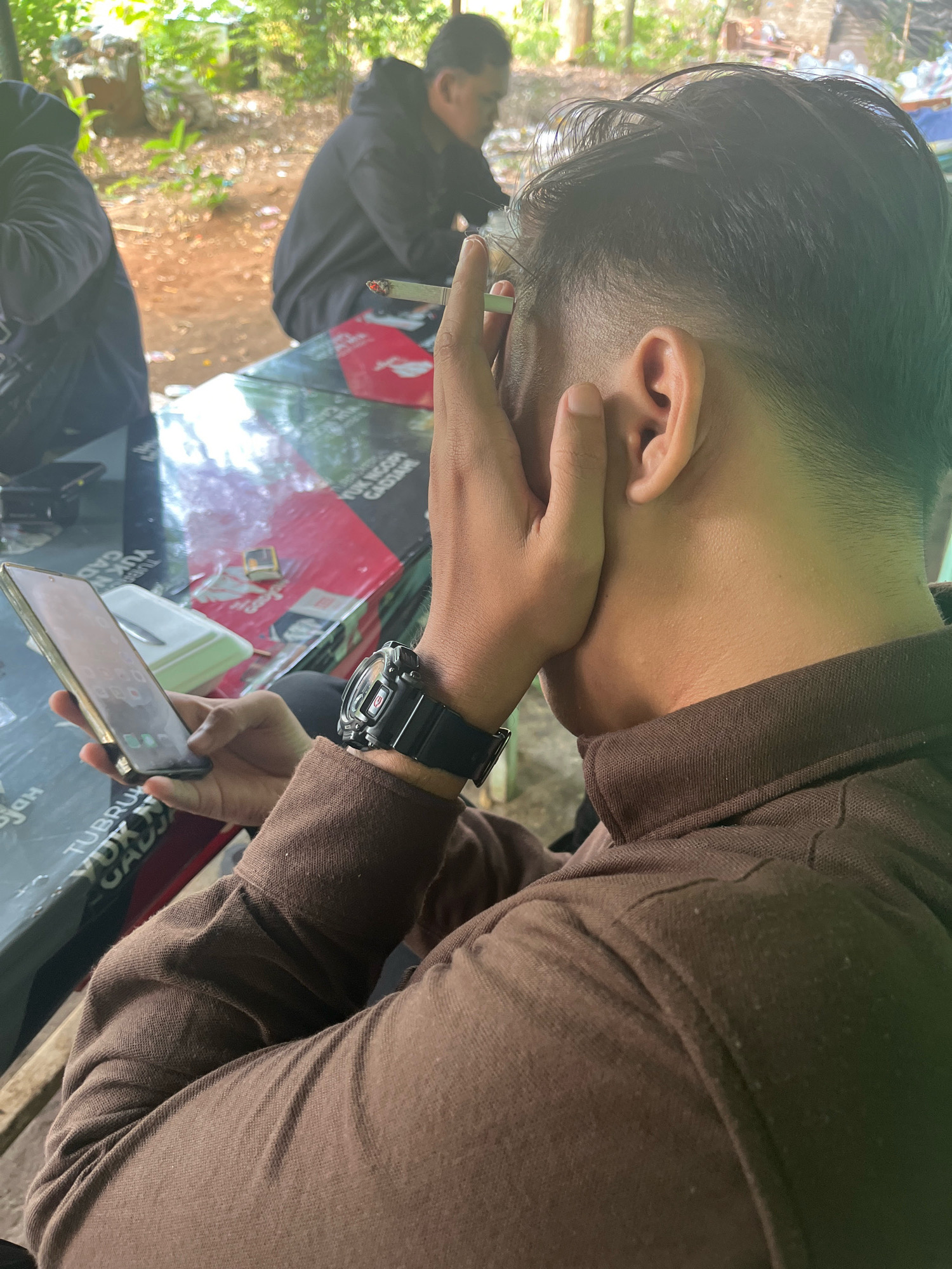 The image captures a group of individuals seated at a table, likely outdoors given the dappled sunlight and lush greenery in the background. The foreground is dominated by a young man wearing a brown hooded jacket, his right hand holding a lit cigarette to his lips. His left hand is partially obscuring his face, and he is looking down at a smartphone he holds. A black digital watch is visible on his left wrist. To the left of the main subject, another person in a black hooded jacket is partially visible. In the background, another individual in a black hooded jacket is also seated at the table. The table itself is covered with various items, including a red and black mat with text and a small white rectangular object. The environment suggests a casual gathering, possibly in a public park or a residential outdoor area. There is no specific indication of the city of Proyonangg, Indonesia.