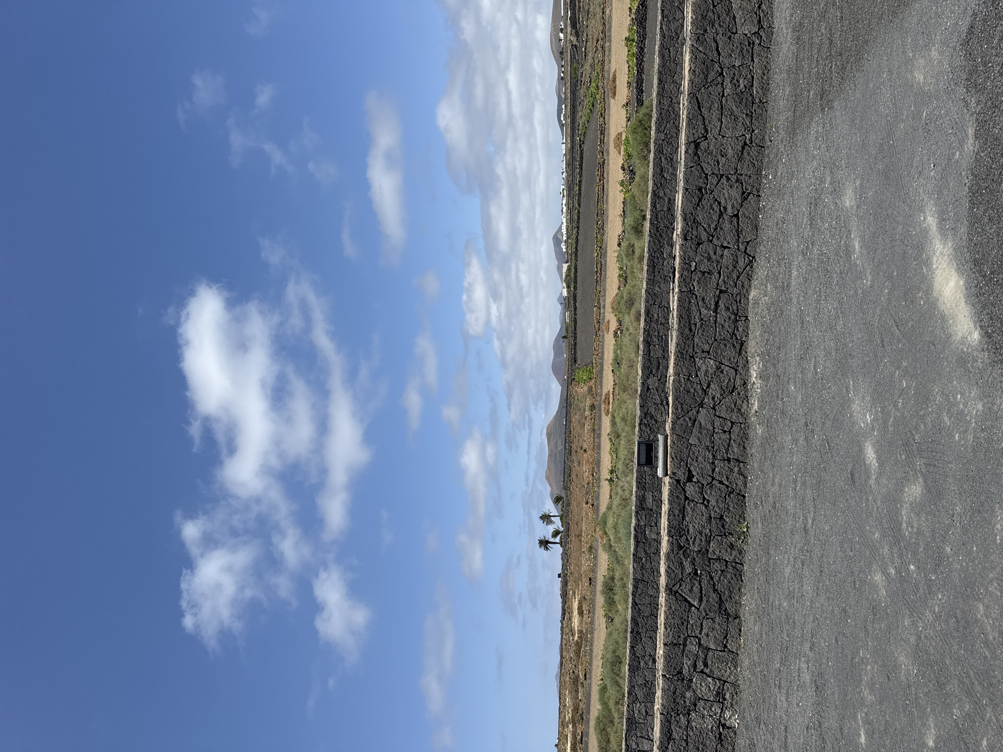 The image depicts a rural landscape in San Bartolomé, Spain. The scene features a dry, arid terrain characterized by dark, volcanic rock walls that define agricultural terraces. These walls, constructed from irregularly shaped dark stones, create a stepped effect across the land.
The sky is a vibrant blue with scattered white clouds, suggesting a sunny day. Vegetation is sparse and primarily consists of dry grass and low-lying shrubs. A few palm trees are visible in the distance, adding a touch of greenery to the otherwise stark environment. In the background, there are hints of mountains and white buildings which is evidence of a town.
The foreground is dominated by a gray, gravelly surface. Affixed to the volcanic rock wall is a square light fixture.