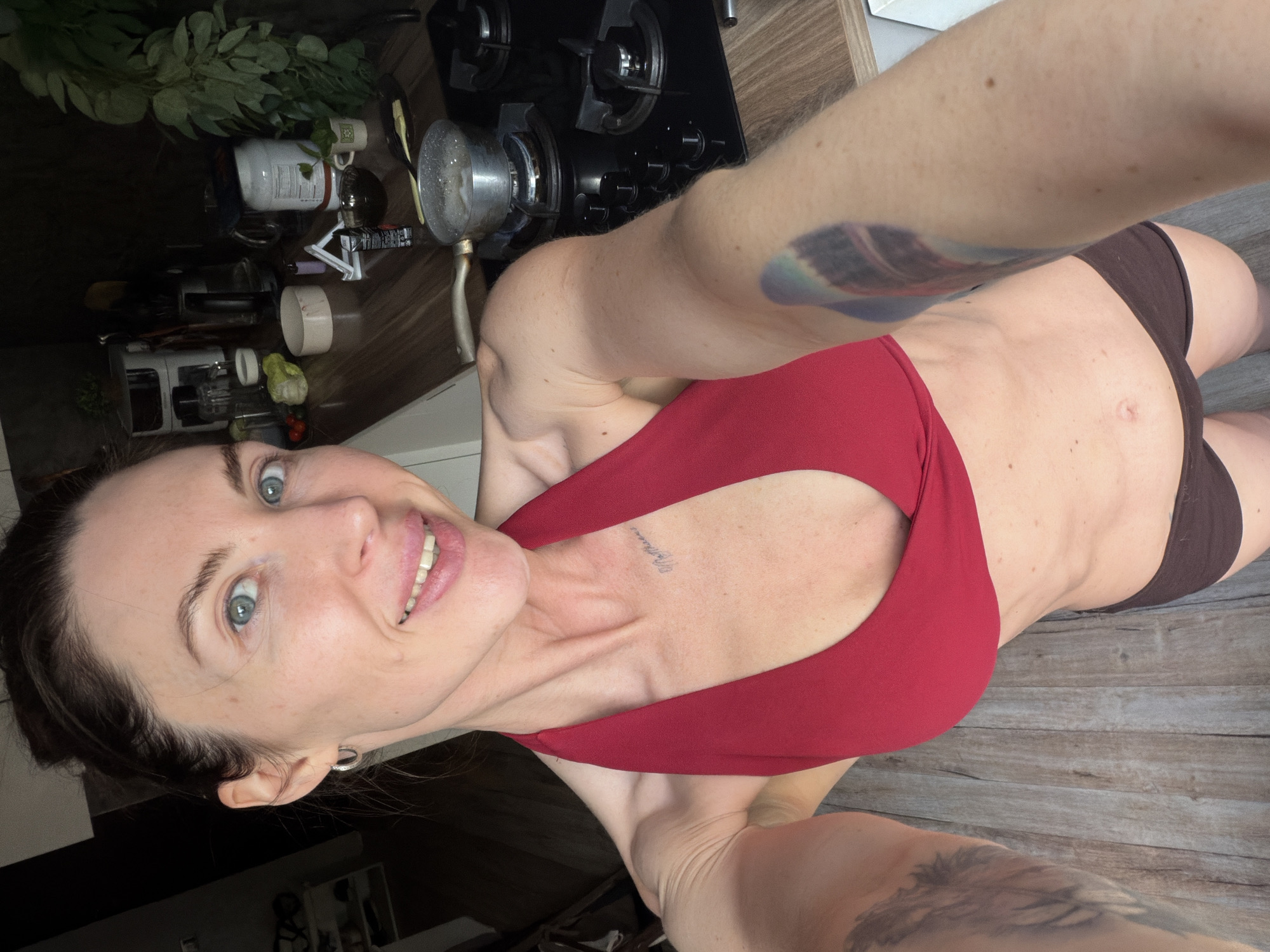 A woman with blue eyes and dark hair, smiling and looking directly at the camera, takes a selfie indoors. She has a fit physique, wearing a red sports bra or top and dark bottoms. A small text tattoo, possibly reading "My Princess" in script, is visible on her upper chest. There is also a larger, colorful tattoo on her upper right arm.
The setting appears to be a kitchen in Perm, Russia, characterized by a wooden countertop and a gas stove with multiple burners. Various kitchen items are scattered on the counter, including a pot with bubbling liquid on one of the burners, suggesting cooking activity. Other visible objects include a blender, several containers, mugs, and what looks like potted greenery or herbs. The floor also has a wooden pattern. The overall lighting suggests an indoor setting, likely during the day. The woman appears cheerful and content.