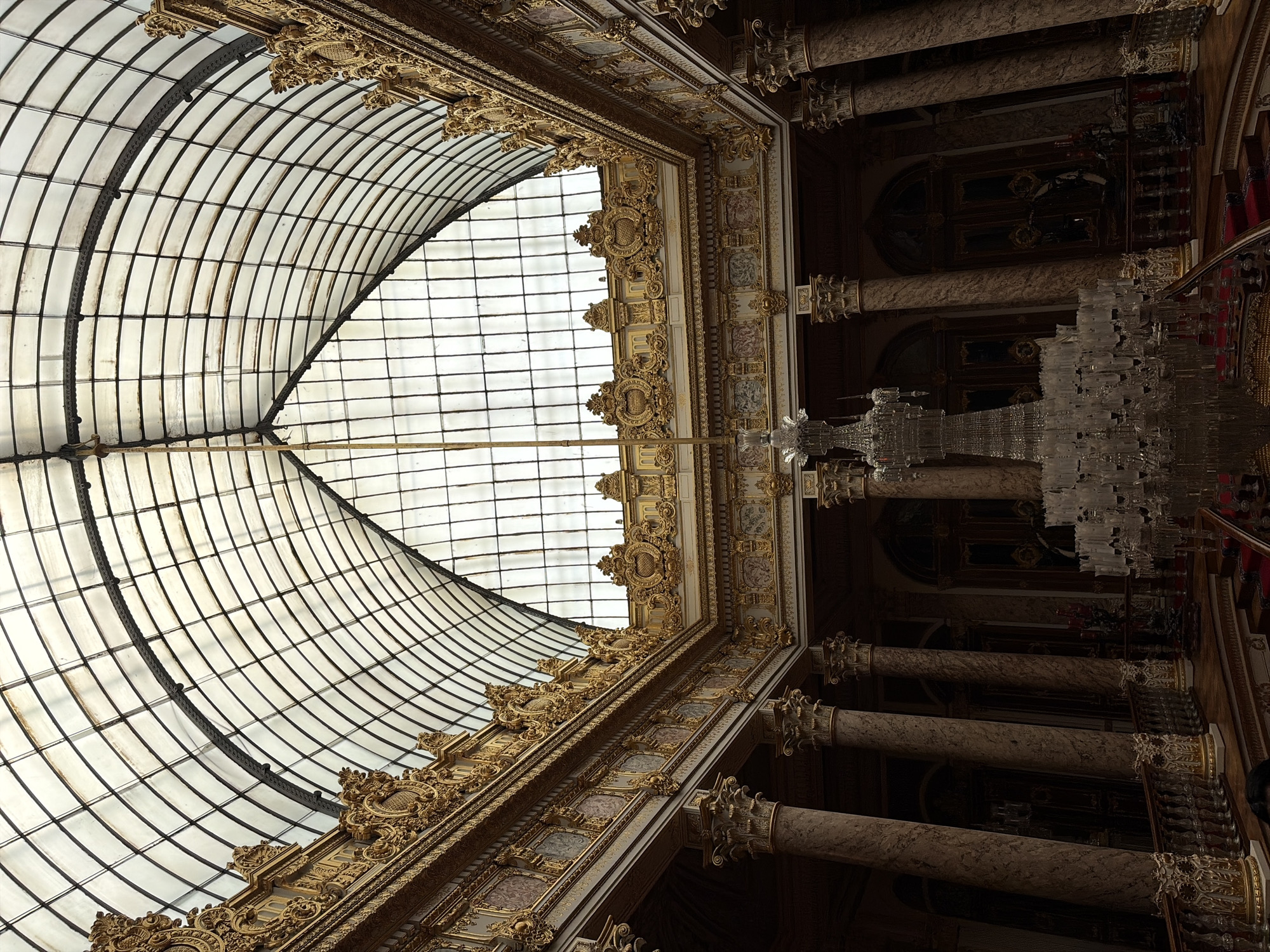 The image is an interior view of Dolmabahçe Palace, located in Beşiktaş, Turkey. The photograph looks upward towards the curved, glass paneled ceiling. Ornate, gold decorations line the edges of the ceiling structure. A large chandelier hangs from the decorated ceiling area. Columns are visible on the right side.