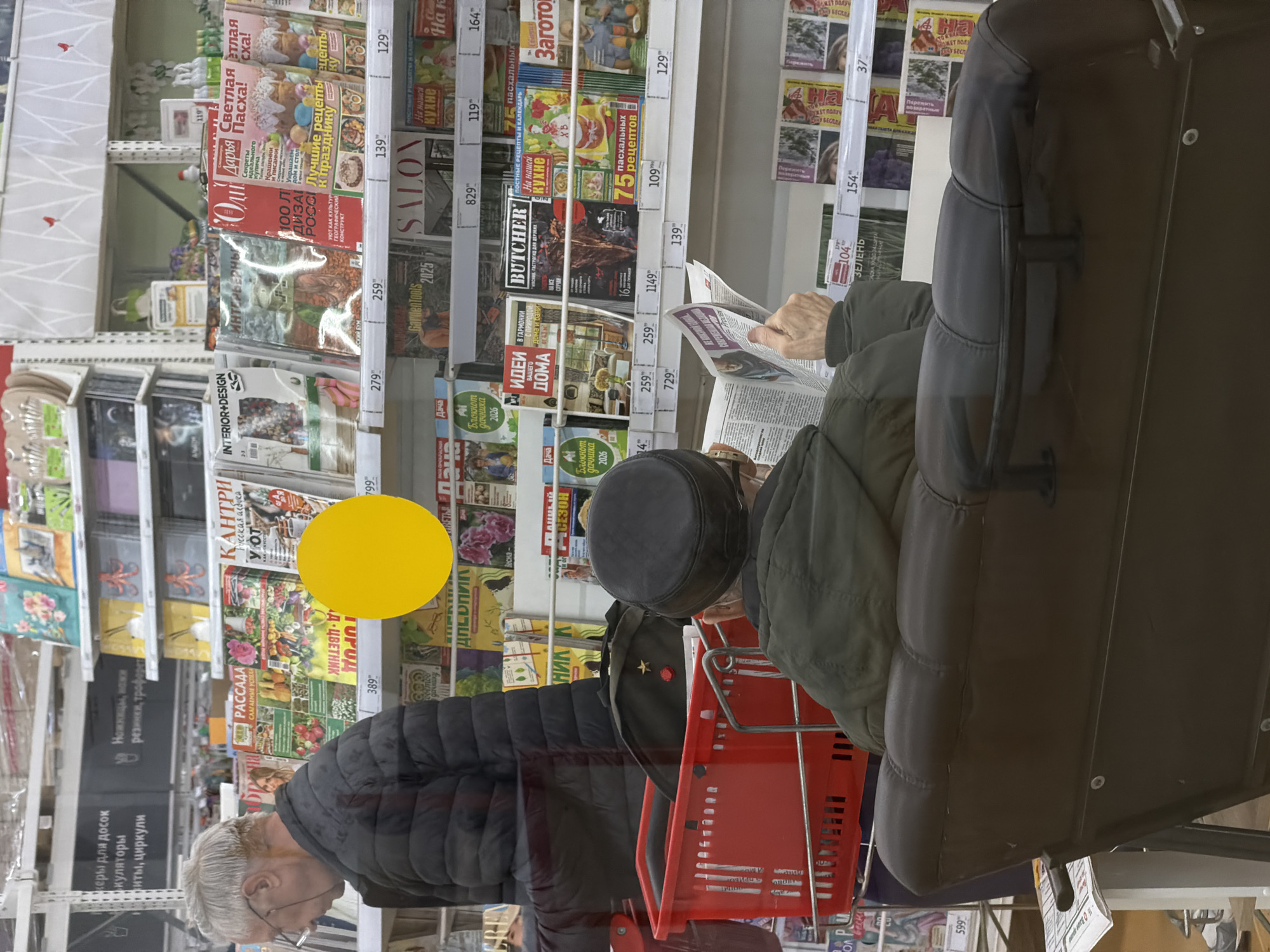 The image shows a scene inside what appears to be a store, possibly in Moscow, Russia, based on the Cyrillic text on the magazine covers. The camera is positioned at an angle, looking toward a magazine rack stocked with various publications on topics such as home and garden, cooking, and lifestyle.
An elderly man with gray hair and glasses is standing in front of the rack. He's wearing a black puffer jacket. Another person in the scene, possibly also male, is wearing a black hat and a green jacket. A red shopping basket is visible, partially obscuring the view of the magazine rack. A third person is seated in a brown chair reading a newspaper.
The shelves are densely packed with magazines, each displaying covers with various designs and titles. Prices are displayed on the shelf edges.
The lighting in the store appears to be fluorescent, common in commercial spaces. The overall impression is that of a routine shopping scene in a well-stocked retail environment.