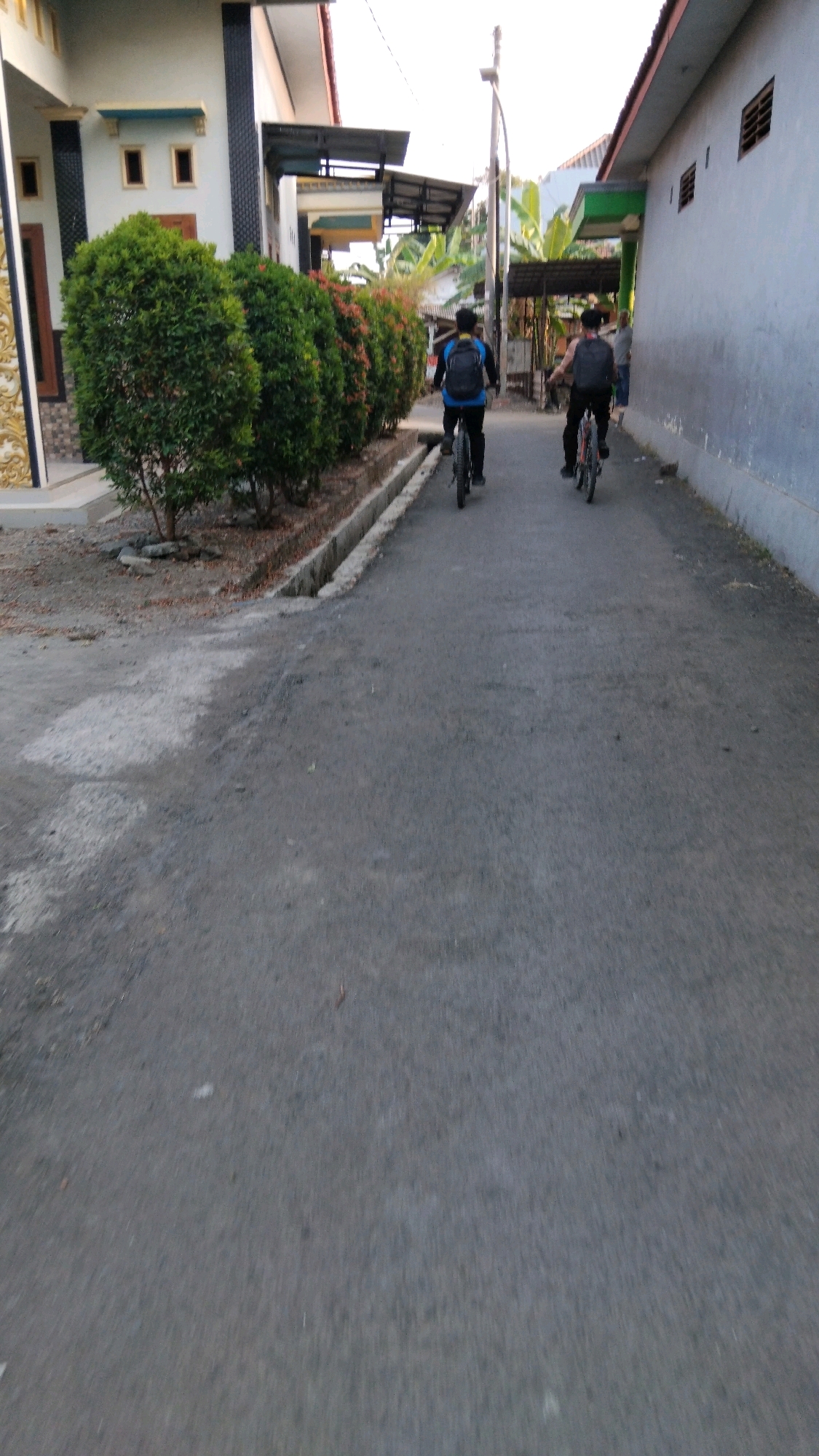The image shows two young men cycling down a paved street in Tegal, Indonesia. The men are wearing backpacks and are on separate bicycles. The street is lined with houses and lush green trees on the left side. The houses are of a simple, modern design with white walls and darker trim. On the right side of the street, there is a house with grey walls and a red-tiled roof. The sky is overcast and the lighting suggests it is late afternoon. The scene appears to be in a residential area with a quiet, everyday atmosphere.