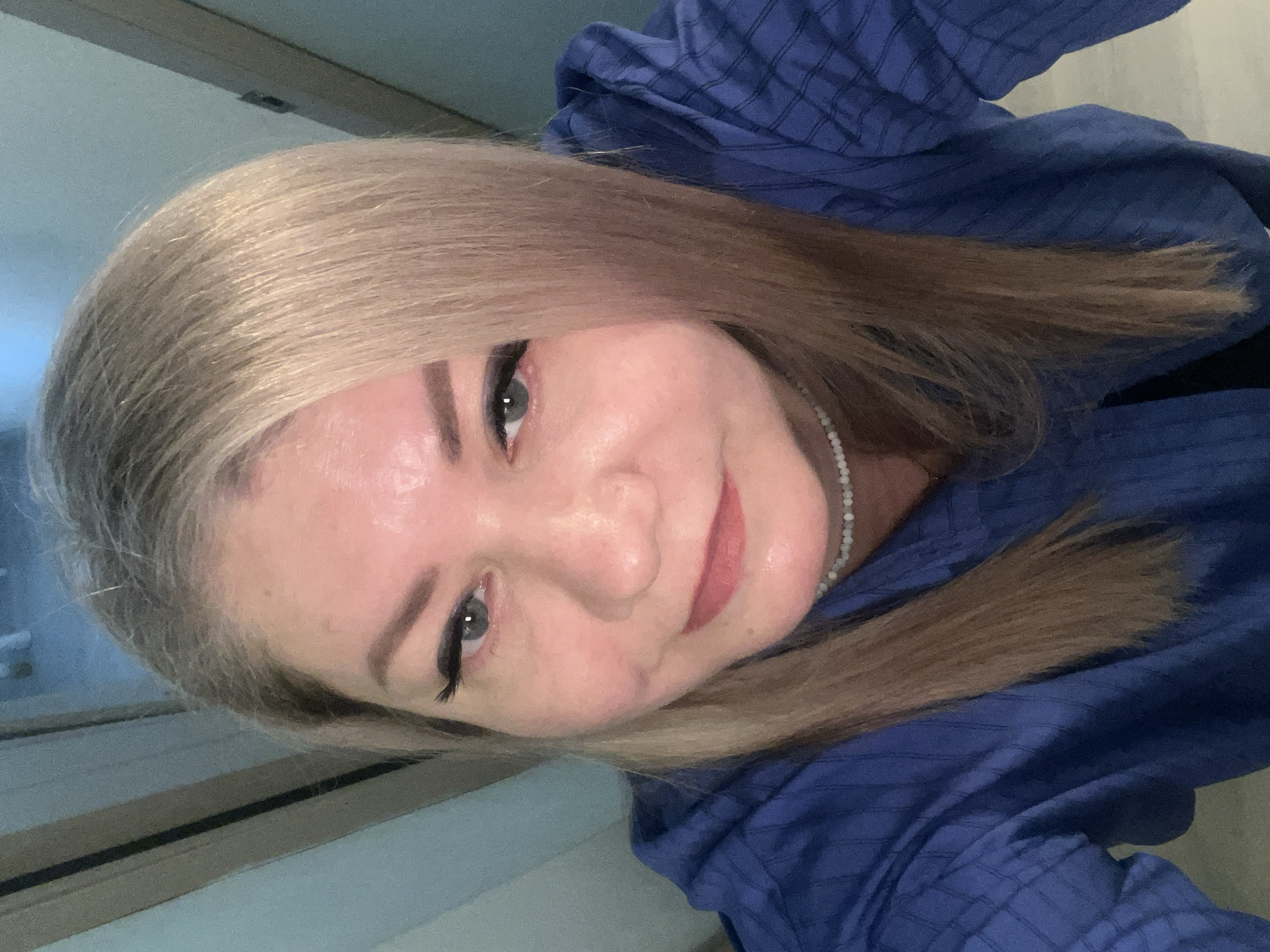 A woman with blonde hair and gray eyes poses for a close-up selfie. She wears a blue patterned shirt and a pearl necklace. The background is nondescript, showing a wall with a window and a door. There is no indication of location in Izhevsk, Russia, or any other specific location.