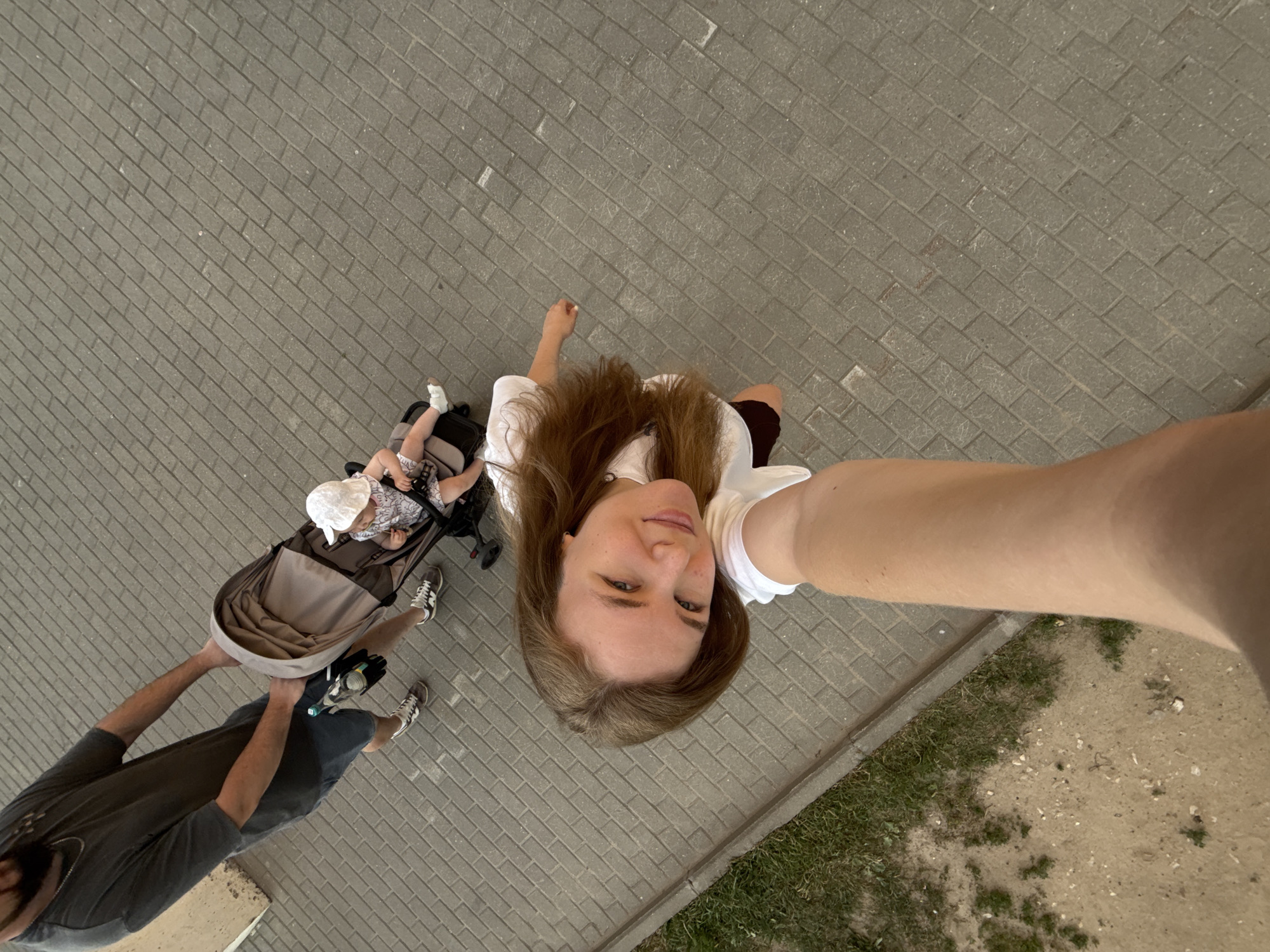 An overhead shot captures a woman taking a selfie as she walks along a paved sidewalk in a city. She is looking towards the camera and smiling. Behind her, a man is pushing a stroller with a baby. The baby is wearing a white hat and is seated in the stroller. The man is wearing a grey t-shirt and shorts. The environment appears to be an urban setting with a grassy area bordering the sidewalk. The paving stones are arranged in a herringbone pattern. The presence of a stroller and a baby suggests a family outing. No specific landmarks are visible to pinpoint the exact location within Vladimir, Russia.