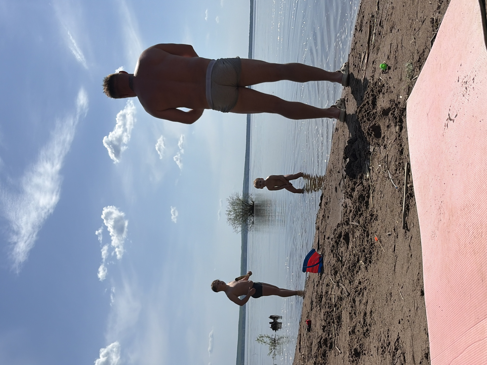 The image depicts a beach scene in Zabegaevo, Russia. There are three people present: a man, a teenager, and a young child. The man is standing near the water's edge, wearing grey briefs. The teenager is holding a bottle, also standing in the water. The child is slightly further out in the water. The environment includes a sandy beach, a body of water, a sky with scattered clouds, and some vegetation visible in the distance. There is a red bucket and a pink pad on the beach. A green ball is also present.