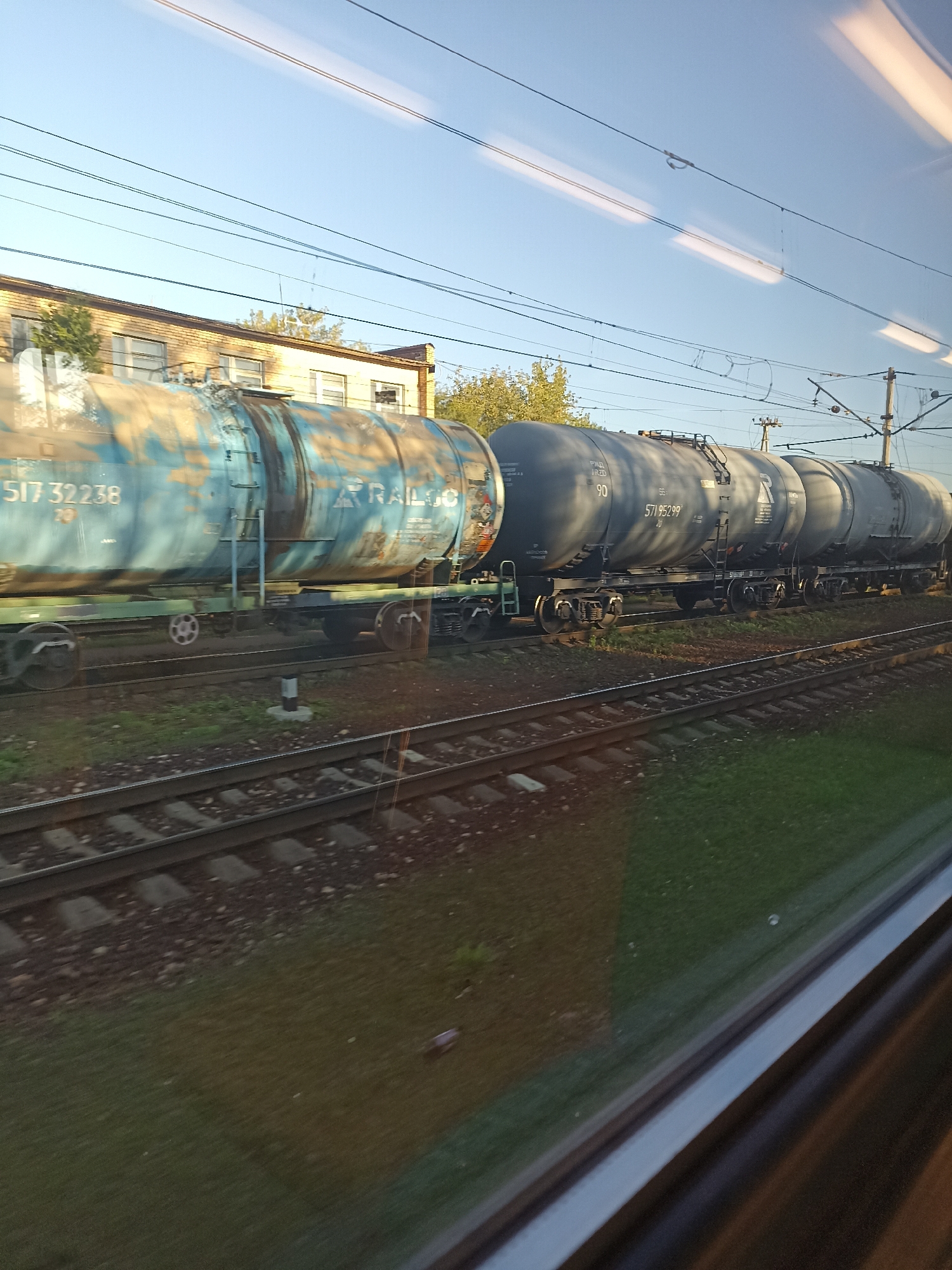 A train of tank cars is seen from the side, moving on a sunny day with a clear blue sky. The train consists of at least two types of tank cars. The first tank car is light blue with significant rust and peeling paint. It has markings including the number "517 32238" and the word "TRAILGO". The second set of tank cars are dark gray and appear to be in better condition. One of these cars has the number "90" and "571 95299" visible. The train is on railroad tracks, with gravel and grass visible along the sides. In the background, there is a light-colored brick building with windows, and some trees. Overhead power lines for the train are also visible. The scene is likely near the city of Podolsk, Russia, given the context of the prompt.