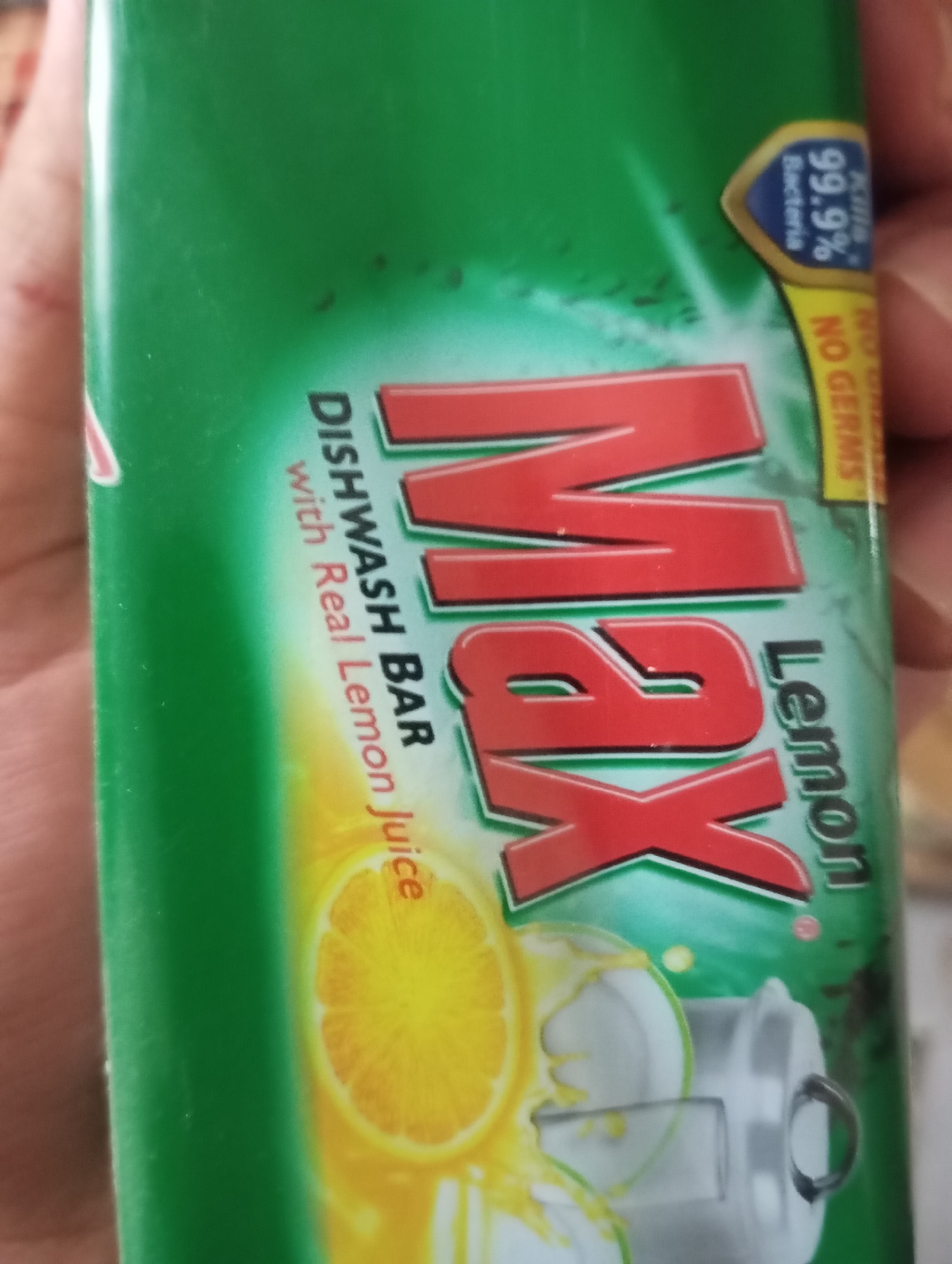 The image shows a close-up of a green package for "Max Dishwash Bar with Real Lemon Juice." The product name "Max" is written in large, red, 3D letters. To the left of "Max," the text "DISHWASH BAR" and "with Real Lemon Juice" is visible. On the right side of the package, the word "Lemon" is written vertically. A stylized image of a lemon slice and suds is present in the lower portion of the package. In the upper right corner, a blue and yellow shield graphic indicates "Kills 99.9% Bacteria" and "NO GERMS." The package is being held by a hand, parts of which are visible on the left and right sides of the image. The background is out of focus and appears to be indoors. There is no discernible information about the location being Hakale, Pakistan.