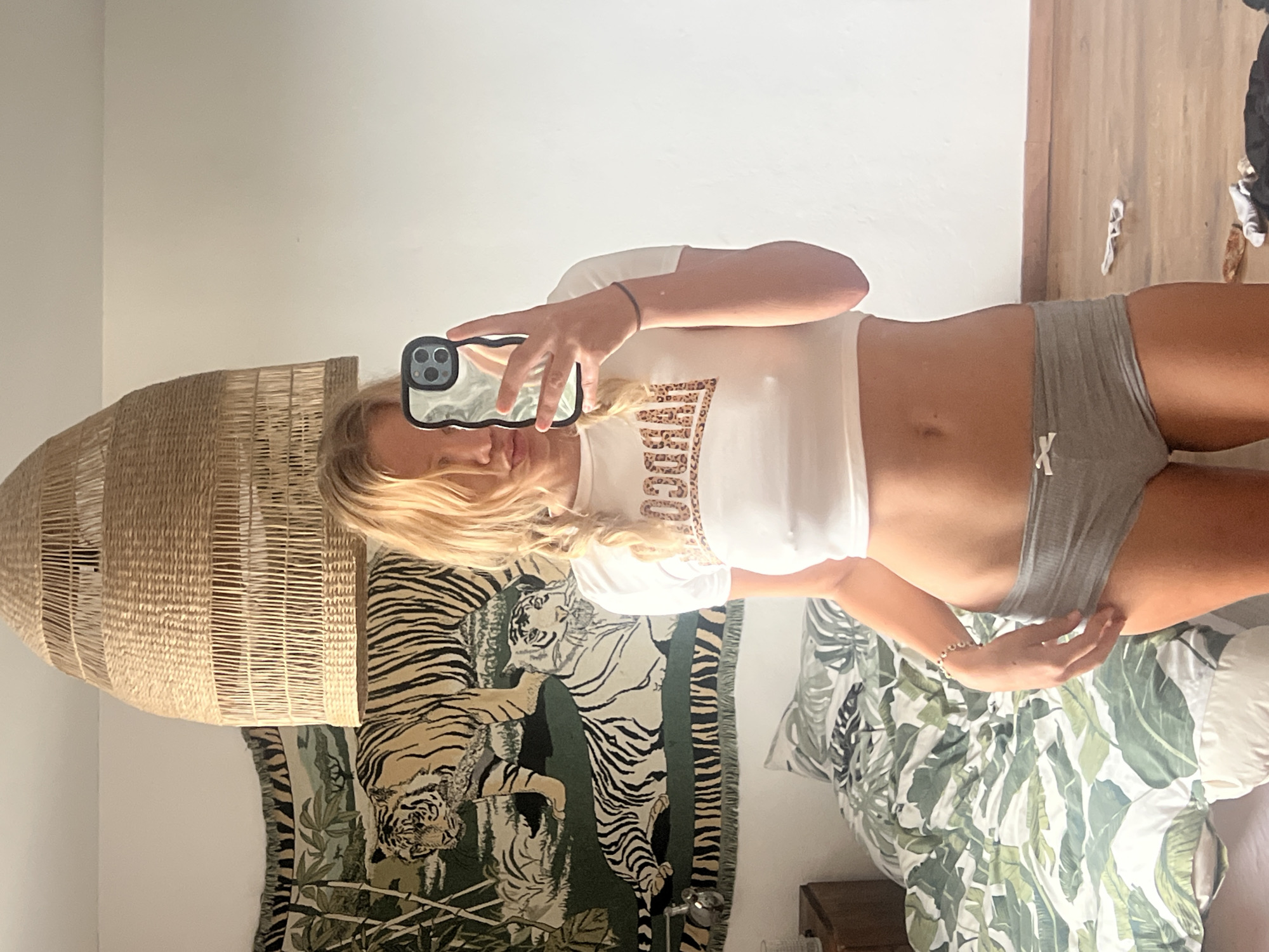 The image shows a young woman taking a mirror selfie in a bedroom. She is wearing a white cropped t-shirt with "JUICY" printed in a leopard print pattern and grey high-waisted shorts. Her blonde hair is in two braids. She has a bracelet on her left wrist and is holding her phone in her right hand, which is reflected in the mirror.
The background of the bedroom includes a large woven pendant light fixture, a tapestry with a tiger print, and a bed with a tropical leaf patterned duvet. The lighting suggests it is daytime. There are no other people visible in the image.