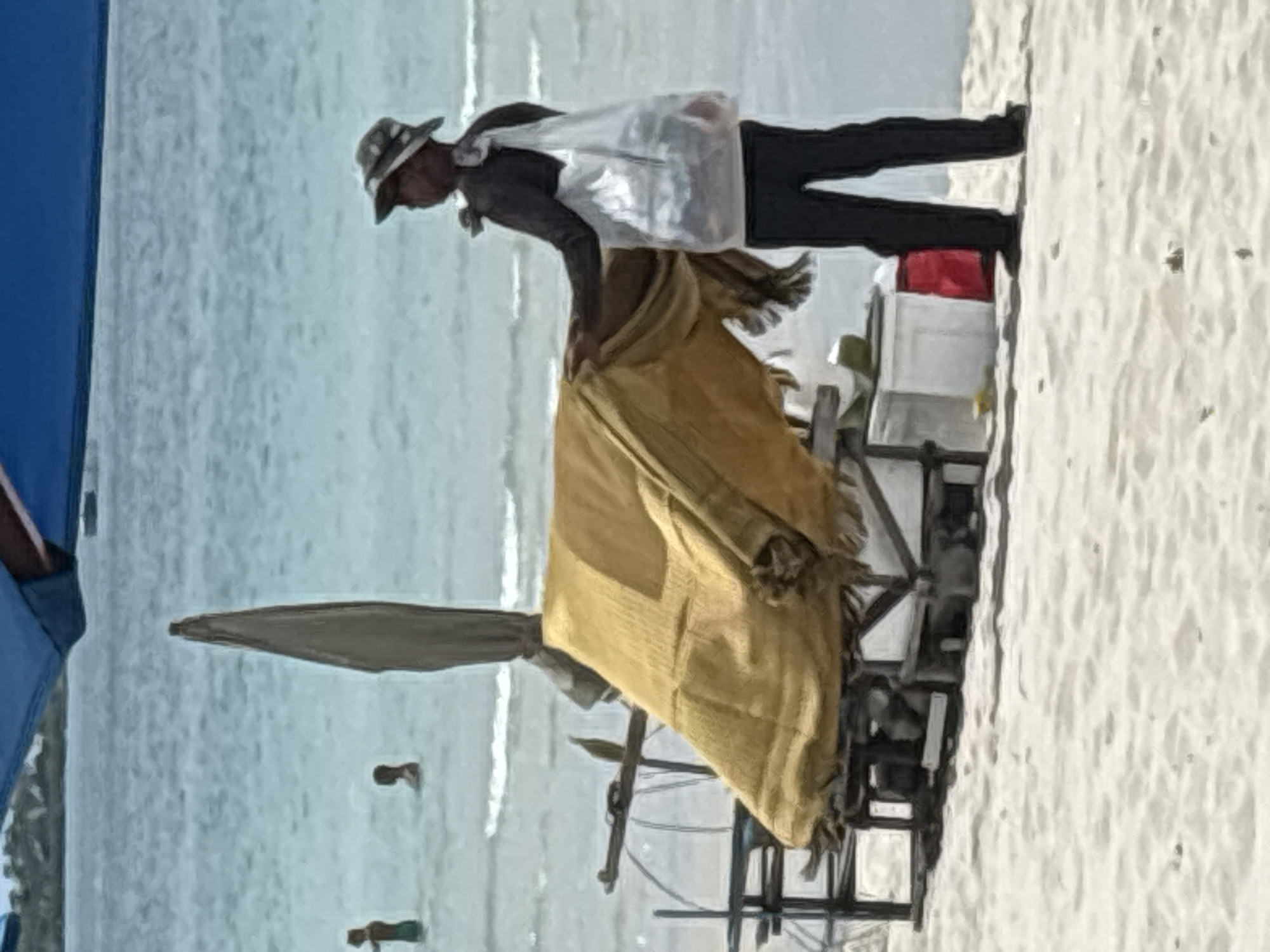 A person wearing a hat and dark clothing stands on a sandy beach in front of a makeshift stall covered with a yellow tarp. The person is holding a translucent bag on their shoulder and appears to be interacting with the stall. Behind the stall, a white cooler and a red item are visible. In the background, the ocean with gentle waves stretches to the horizon, and two individuals are seen in the water. The scene is framed by a portion of a blue umbrella.