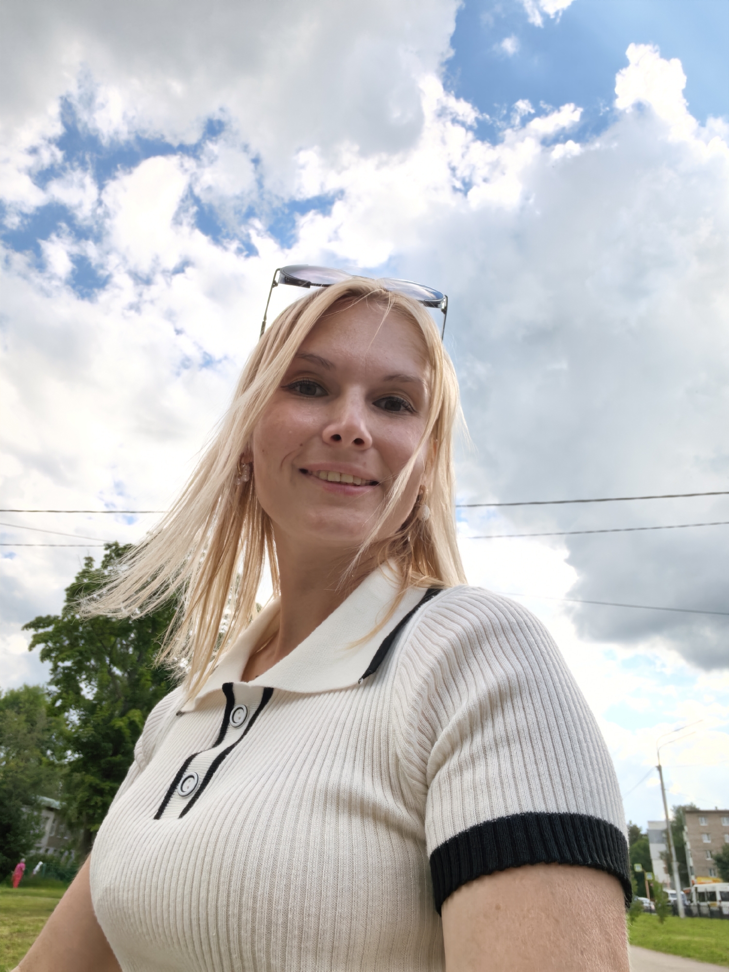 A young woman with blonde hair and sunglasses pushed up on her head is looking at the camera from a low angle. She is wearing a white, ribbed polo shirt with black trim and buttons. The background is a cloudy sky with trees and buildings visible to the left and right. The image was taken outside in an urban environment, likely in Sterlitamak, Russia, as suggested by the presence of typical Russian residential buildings and a bus in the distance.