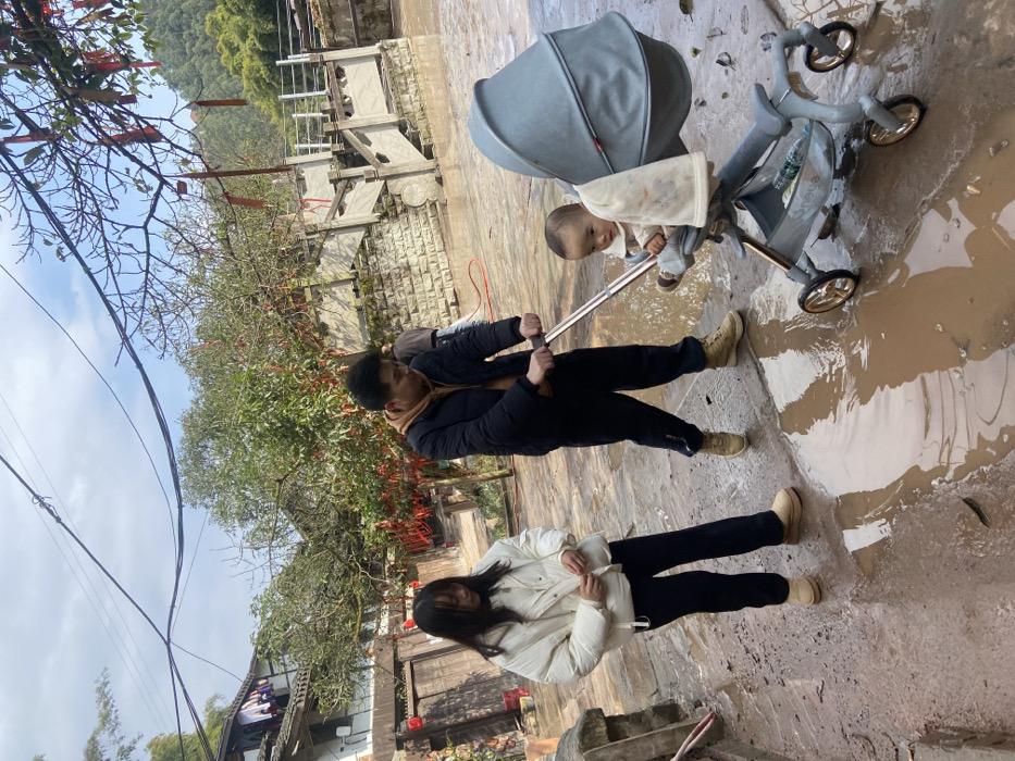 A family of three on a paved surface with puddles is visible. A man in a black jacket pushes a stroller containing a baby. A woman wearing a white jacket stands near the stroller. Trees are in the background along with buildings. Overhead power lines are visible, as well as trees with decorative red strips hanging from them.