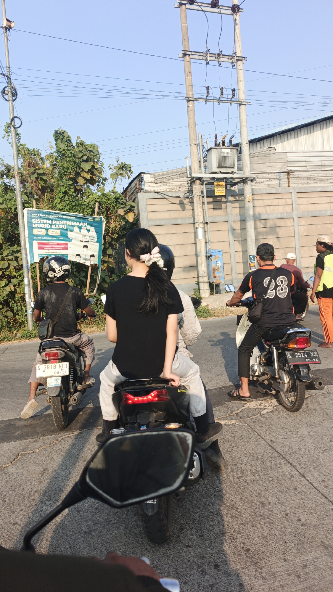 The image depicts a street scene in Sumolawang, Indonesia, under a clear sky. The foreground shows the handlebars and a portion of the mirror of a motorcycle, suggesting the image is taken from the perspective of a rider. Following the road, several people on motorcycles are visible.
In the middle ground, three motorcycles are moving. The closest one has a rider with dark hair and a light-colored scrunchie holding their ponytail back, wearing a black t-shirt and light grey pants. They are seated behind another rider wearing a black helmet. To their left, another motorcycle carries a rider wearing a black t-shirt with the number "28" in white on the back and a black cap, paired with black pants and black sandals. This rider is also followed by a passenger. Further to the left, a motorcycle has a rider wearing a black t-shirt and grey pants, with a black helmet.
In the background, a large electrical transformer is mounted on a utility pole, with numerous wires stretching across the frame. A light-colored building with a textured wall runs along the right side of the road, and to the left, lush green foliage surrounds a large sign. The sign, partially obscured by trees, displays text in Indonesian, including "SMP & SMA AL-QUR'AN KHASNAWI" and information about new student enrollment.
The road surface is paved with grey concrete, showing visible expansion joints and tire marks. The overall scene captures a typical moment of daily transportation in this Indonesian town.