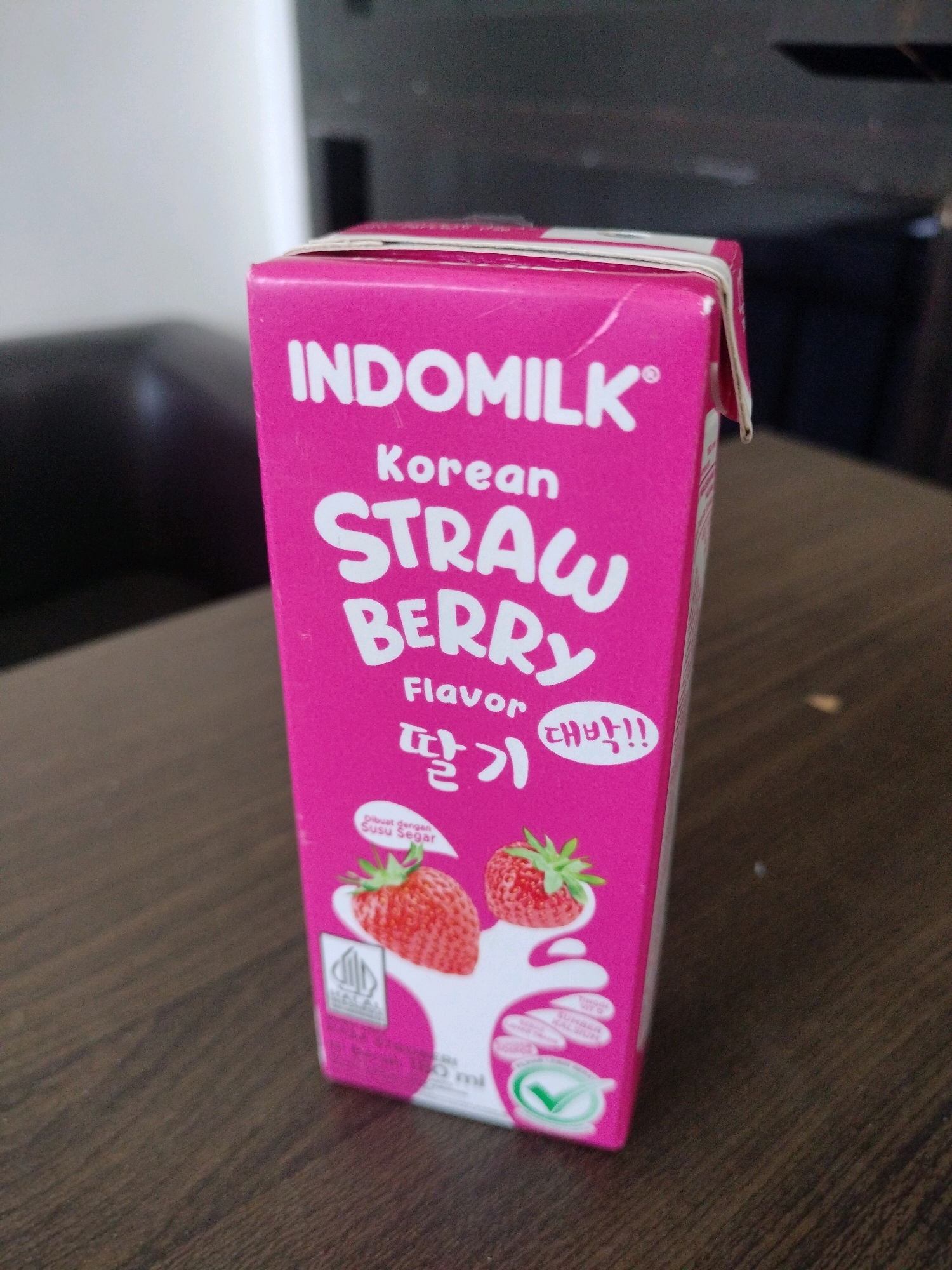A pink carton of Indomilk Korean Strawberry Flavor milk is positioned on a dark wood table. The carton features prominent white lettering that reads "INDOMILK," "KOREAN," "STRAW BERRY," and "Flavor." Below the English text, Korean characters are visible, and a smaller text bubble says "대박!!". Images of strawberries and milk splashes are printed on the carton, along with a halal logo and a green checkmark symbol. The carton is secured with a white rubber band. The background is blurred, showing dark furniture and a white wall. The lighting suggests an indoor environment. No people are visible in the image, and no specific details about the location in Kel. Karang Pamulang, Indonesia are provided within the image itself.