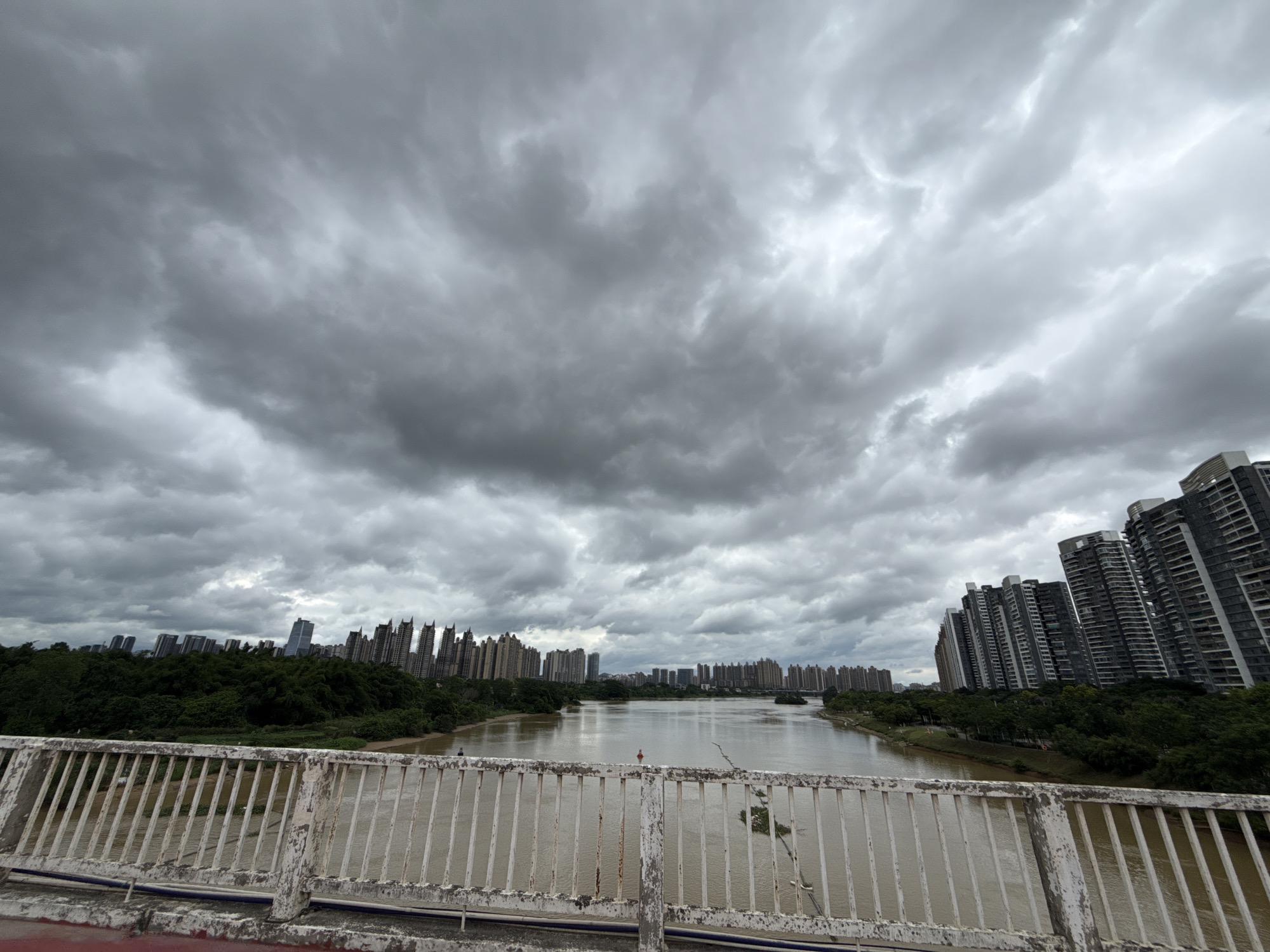 The image shows a wide, muddy river flowing through a city under a cloudy sky. The foreground features a white metal bridge railing, which appears weathered and somewhat rusted. Beyond the bridge, the river stretches into the distance, bordered by dense green trees on the left bank and a series of tall, modern apartment buildings on the right bank. The buildings are varied in design, with some appearing sleek and contemporary, while others have a more angular, stepped facade. The sky is filled with heavy, gray clouds, suggesting overcast weather or an approaching storm. The overall atmosphere is somewhat somber due to the muted light and the imposing cloud cover. The scene depicts an urban environment in Huizhou, China, likely a residential area with a riverfront developed with high-rise buildings and green spaces. No people are clearly visible in the scene, though there might be distant figures on the river or within the buildings.