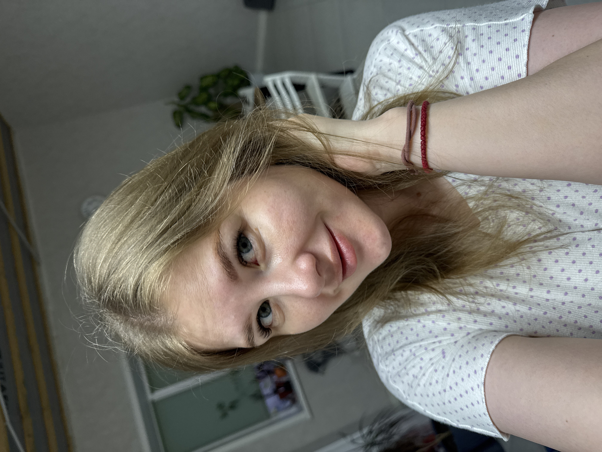 A woman with blonde hair and blue eyes is indoors, facing the camera with a slight smile. She is wearing a white, long-sleeved shirt with small purple polka dots. On her left wrist, she is wearing two bracelets: one is a red beaded bracelet and the other is a tan-colored woven bracelet. A plant and a white chair are visible in the background, and a framed picture hangs on the wall. The location is Gornyi (Dvurechenskoe S/P), Russia.