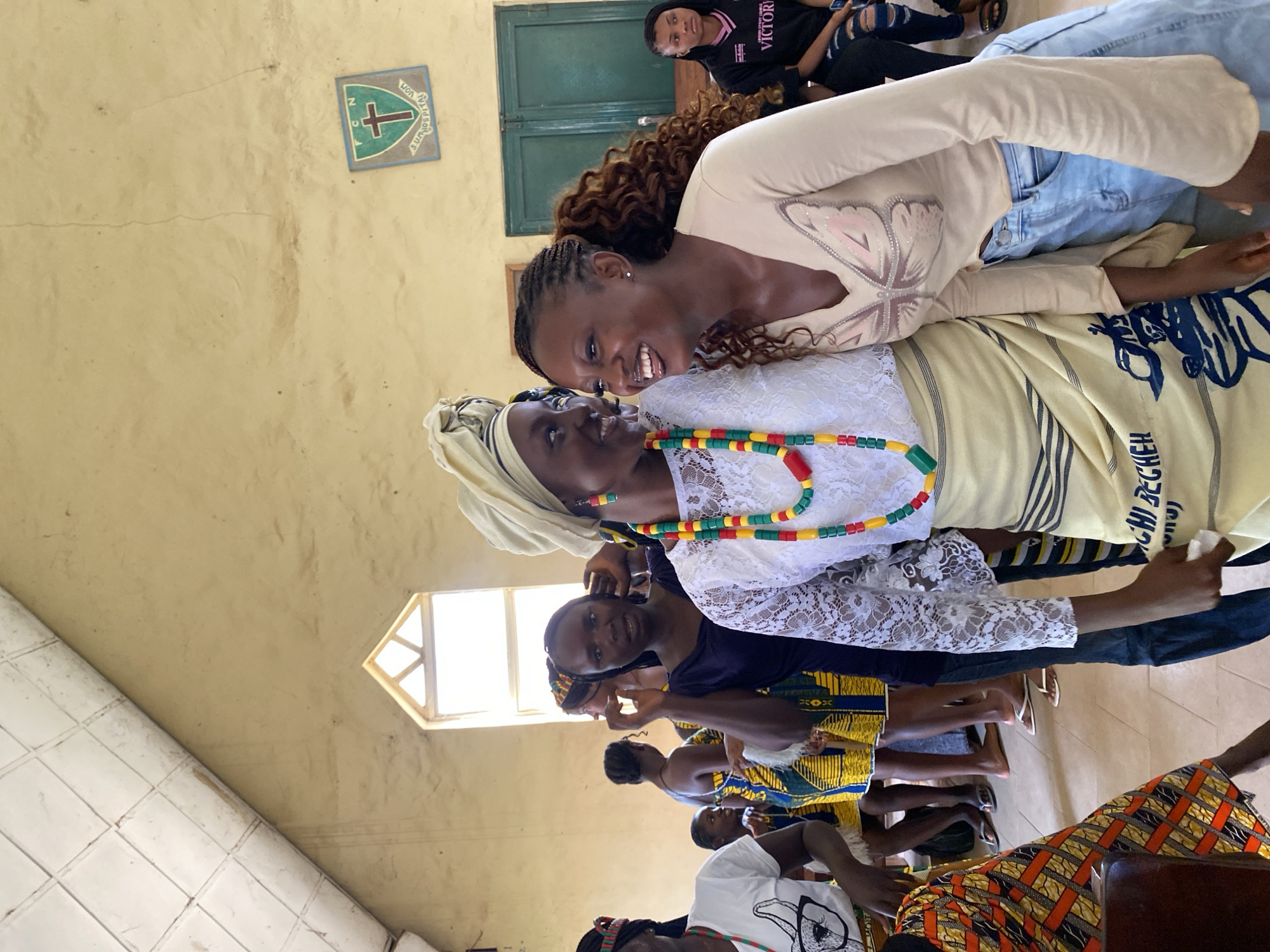 The image depicts a gathering of people, primarily women, in an indoor setting that appears to be a community hall or church in Dutsen Kato, Nigeria. The room has pale yellow walls and a tiled floor. A small, diamond-shaped window with a light-colored frame is visible, suggesting natural light entering the space. There is a green window with multiple panes above a person seated in the background. On the wall, a square emblem with a cross and some text is displayed.
Several women are present, some facing the camera and others turned away. The woman in the foreground is leaning in to speak to an older woman, and both are smiling, conveying a sense of warmth and connection. The older woman is wearing a headscarf and a white lacy top, adorned with a colorful beaded necklace featuring red, yellow, green, and black beads. The younger woman is wearing a light-colored long-sleeved shirt with a butterfly design on it and jeans. Other women in the background are dressed in vibrant, patterned African attire. The overall mood seems joyful and social, possibly a celebration or a community meeting.
The image is oriented vertically, suggesting it might have been taken in portrait mode or that the camera was held at an angle. The lighting is somewhat uneven, with some areas brighter than others. The time of day appears to be daytime due to the natural light from the window. Visible text includes "VICTORIA" on a black t-shirt worn by a person in the background, and partial text on the beaded necklace and on the emblem on the wall, which is difficult to decipher clearly. The presence of traditional clothing and the general atmosphere suggest a cultural or religious event.