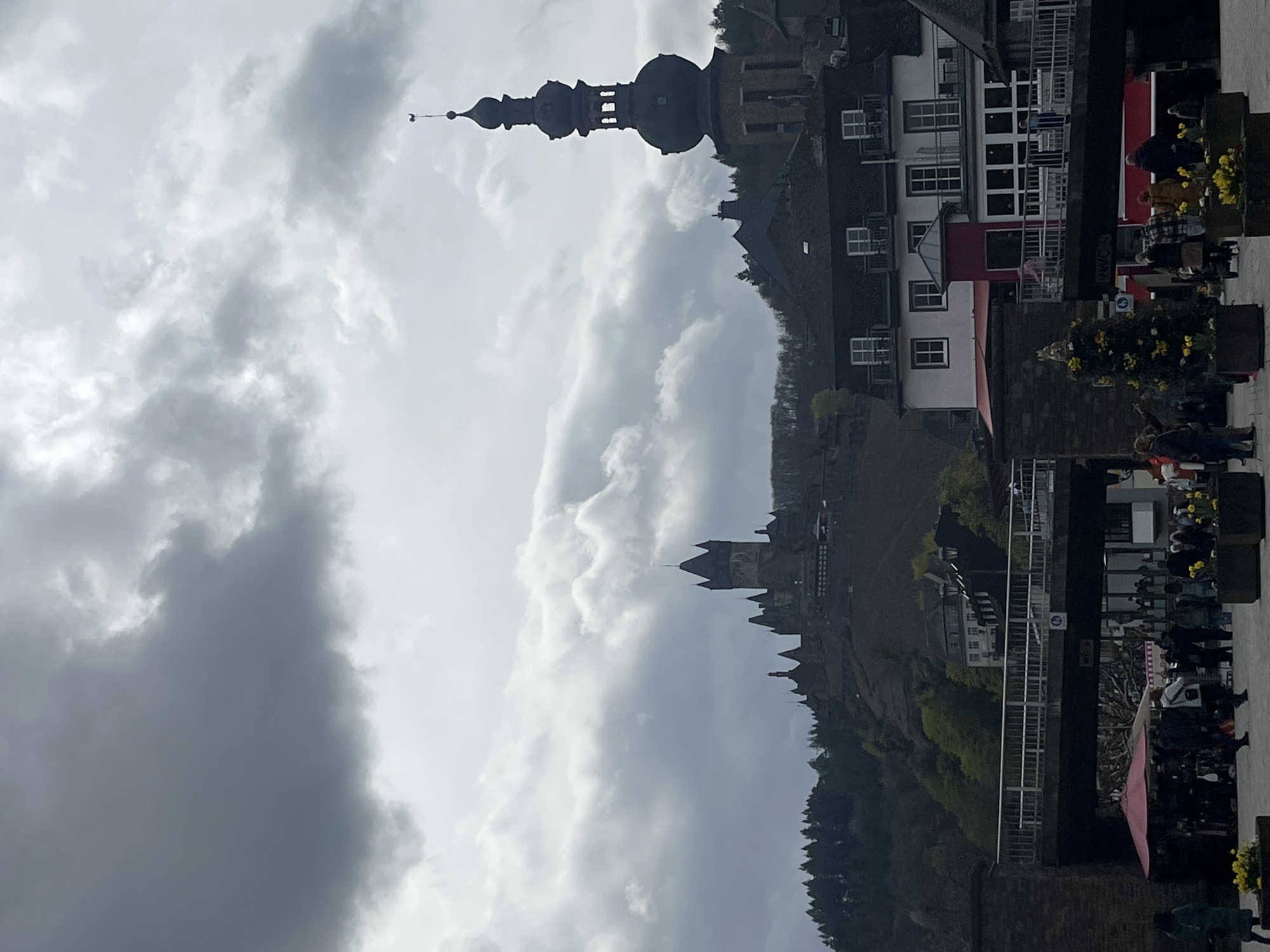 This image depicts Cochem, Germany. The sky is overcast with heavy clouds. A tall, historic church steeple with multiple domes dominates the view. A river flows through the center of the frame. Buildings line the riverbank with classic European architecture, featuring white and red facades. People are seen walking along a paved walkway near the river. They are blurred, suggesting motion, and some are interacting in small groups. Flower planters with yellow flowers line the walkway, adding color. Dense, green foliage covers a hillside, extending from the river up to the buildings. A bridge crosses the river, connecting the two sides of Cochem. The photo has a slightly rotated orientation.