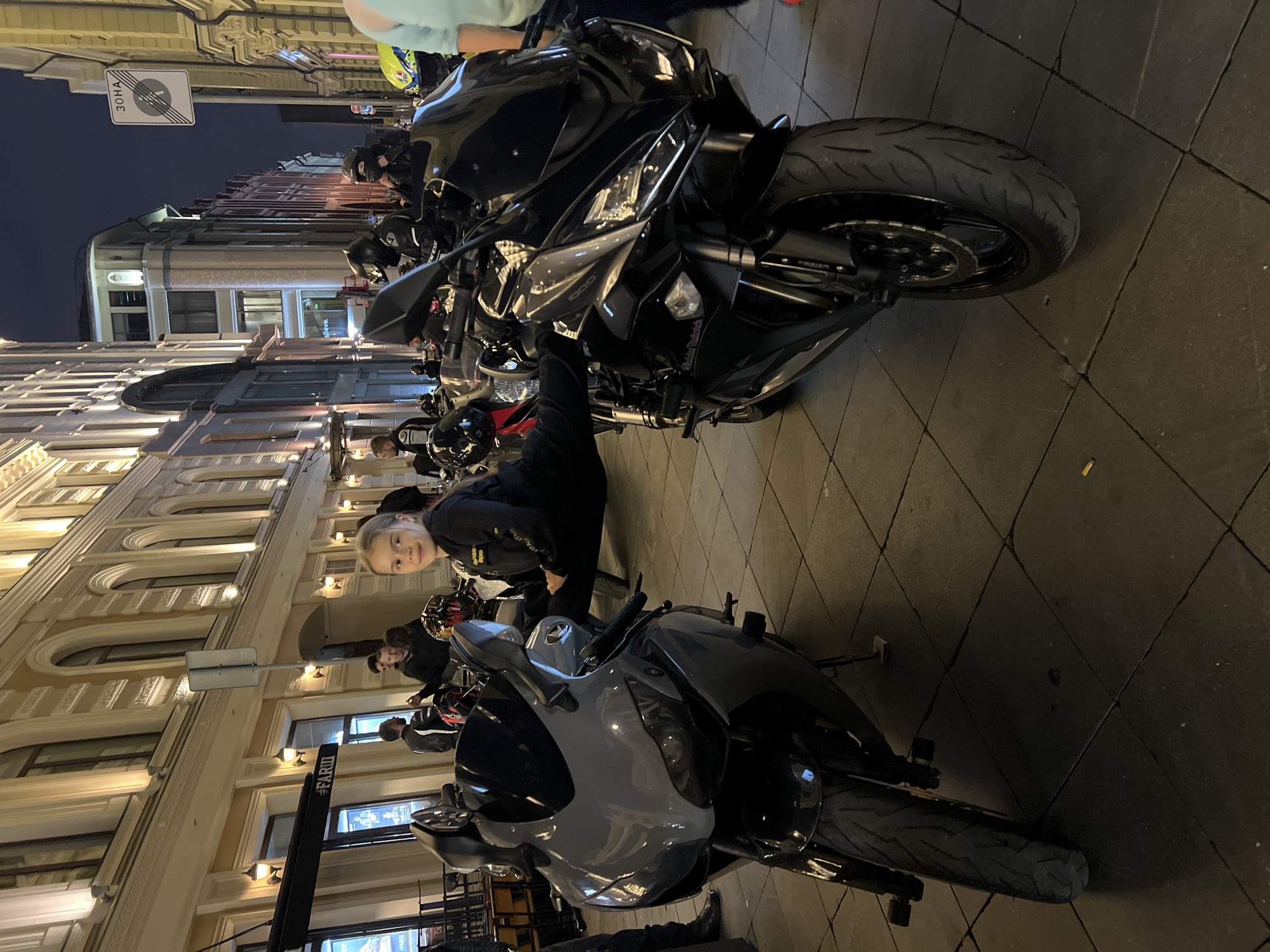 A group of people stand around several parked motorcycles on a city street at night. The scene appears to be in Moscow, Russia, based on a sign with Cyrillic text visible in the background. The street is paved with dark square tiles, and ornate buildings with illuminated windows line the street. A few streetlights cast a warm glow on the scene, suggesting it is evening or night. The people are dressed casually, and some are wearing motorcycle helmets. One young woman in the foreground is smiling at the camera, leaning against a black motorcycle. The motorcycles are modern sportbikes, some black and one a lighter gray. The atmosphere seems relaxed and social, with people gathering around the bikes. A sign with the Cyrillic word "ЗОНА" (ZONA), meaning "ZONE," is visible on a pole.