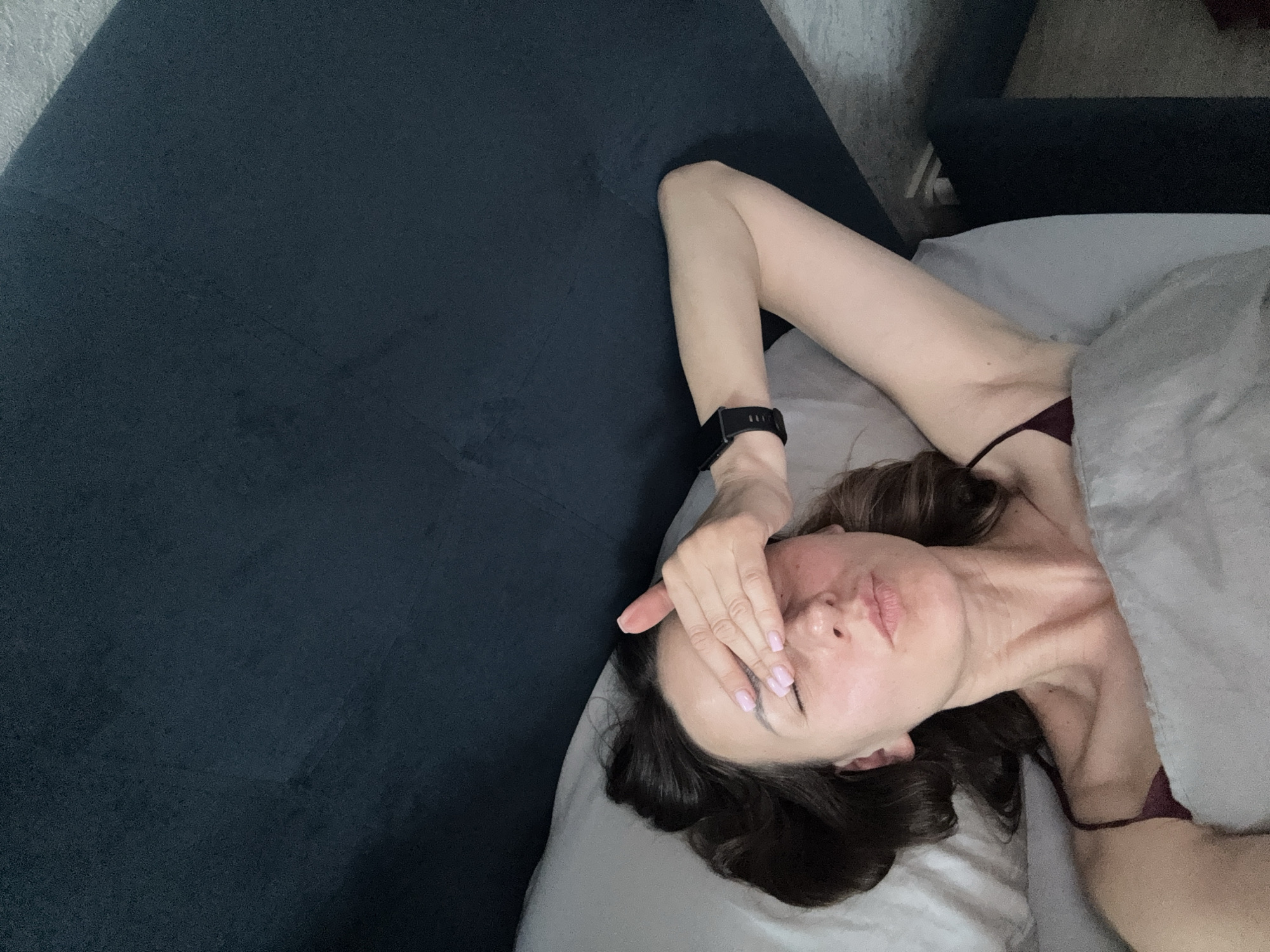 A high-angle selfie captures a woman lying in a bed indoors. The woman has dark brown hair and is positioned on her back with her head resting on a white pillow. Her right hand, adorned with light pink nail polish, covers her eyes, with fingers extending across her forehead. Her lips are slightly pursed. She is wearing a maroon-colored spaghetti strap top. A dark digital watch is present on her left wrist. A light grey blanket covers a portion of her body. The headboard of the bed behind her is a dark blue or deep teal, textured fabric. To the right of the bed, a light-colored wall or curtain is partially visible, along with a dark object that appears to be furniture and a lighter object on the floor. The image contains no discernible visual elements to indicate the specific location as the city of Sochi, Russia.