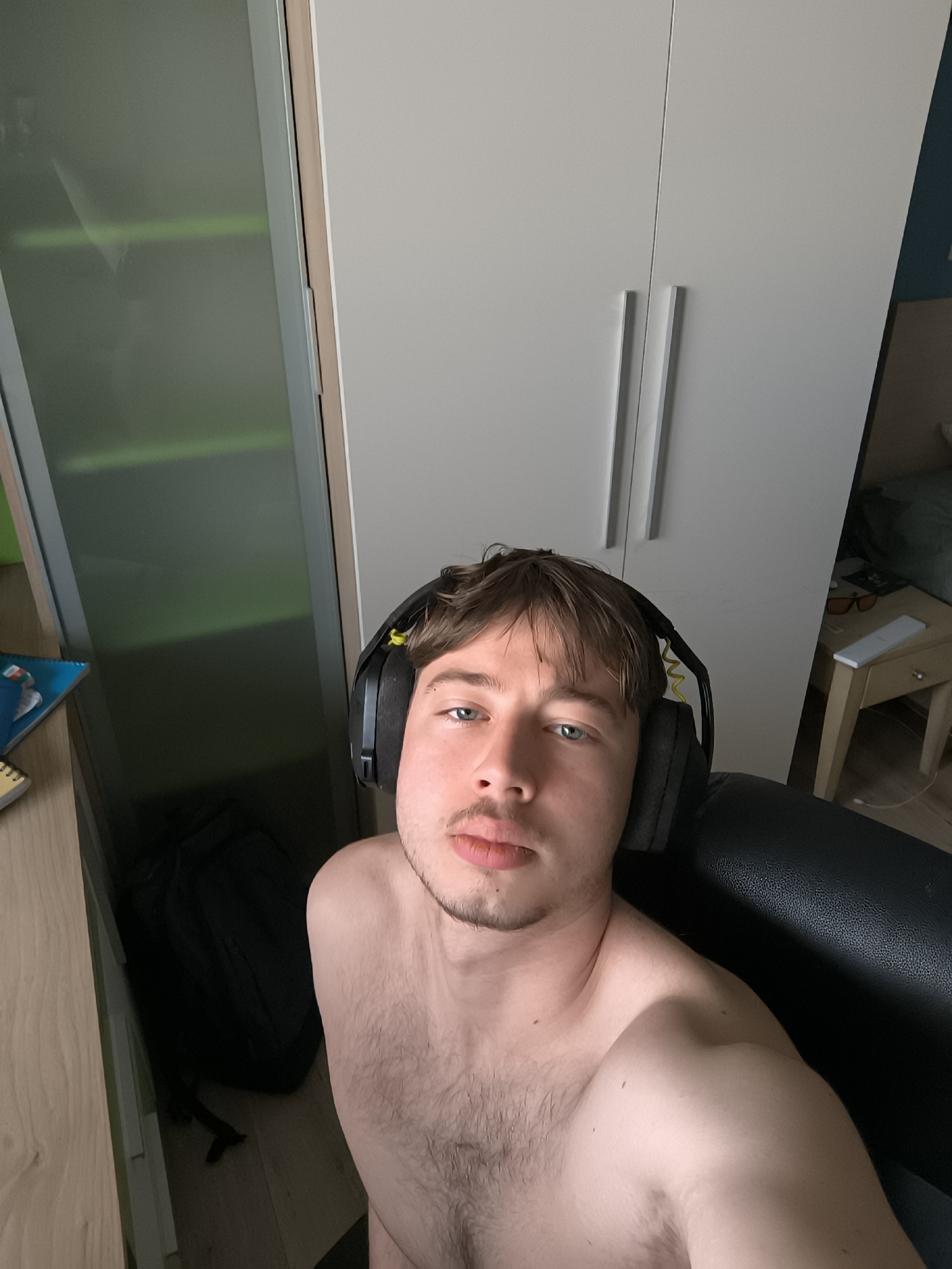 A shirtless man with short brown hair and a stubble beard is centered in the frame, wearing black headphones. He is seated in a black chair. Behind him is a wardrobe with white doors and silver handles, and to the left a mirrored door and a desk. A black backpack is visible on the floor near the desk. A bedside table with items is also visible in the background. The image suggests an indoor setting, likely a room in an apartment or house. There is no indication that the photo was taken in Sofia, Bulgaria.