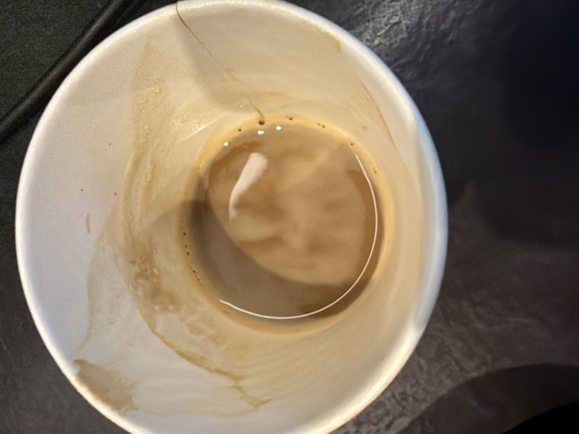 The image provides an overhead view of an off-white paper cup containing a small amount of light brown liquid, presumably coffee with milk. The inner walls of the cup show dried streaks and residue from the beverage, indicating it has been partially consumed. A vertical seam or fold line is visible near the top right rim of the cup.
On the surface of the remaining liquid, there are small bubbles or crema, and a prominent, curved white reflection suggests an overhead light source. The cup rests on a dark, textured surface, possibly a table, which appears to be indoors. There are no people present, nor are there any discernible activities taking place beyond the implied consumption of the drink. No text is visible on the cup or in the background.
