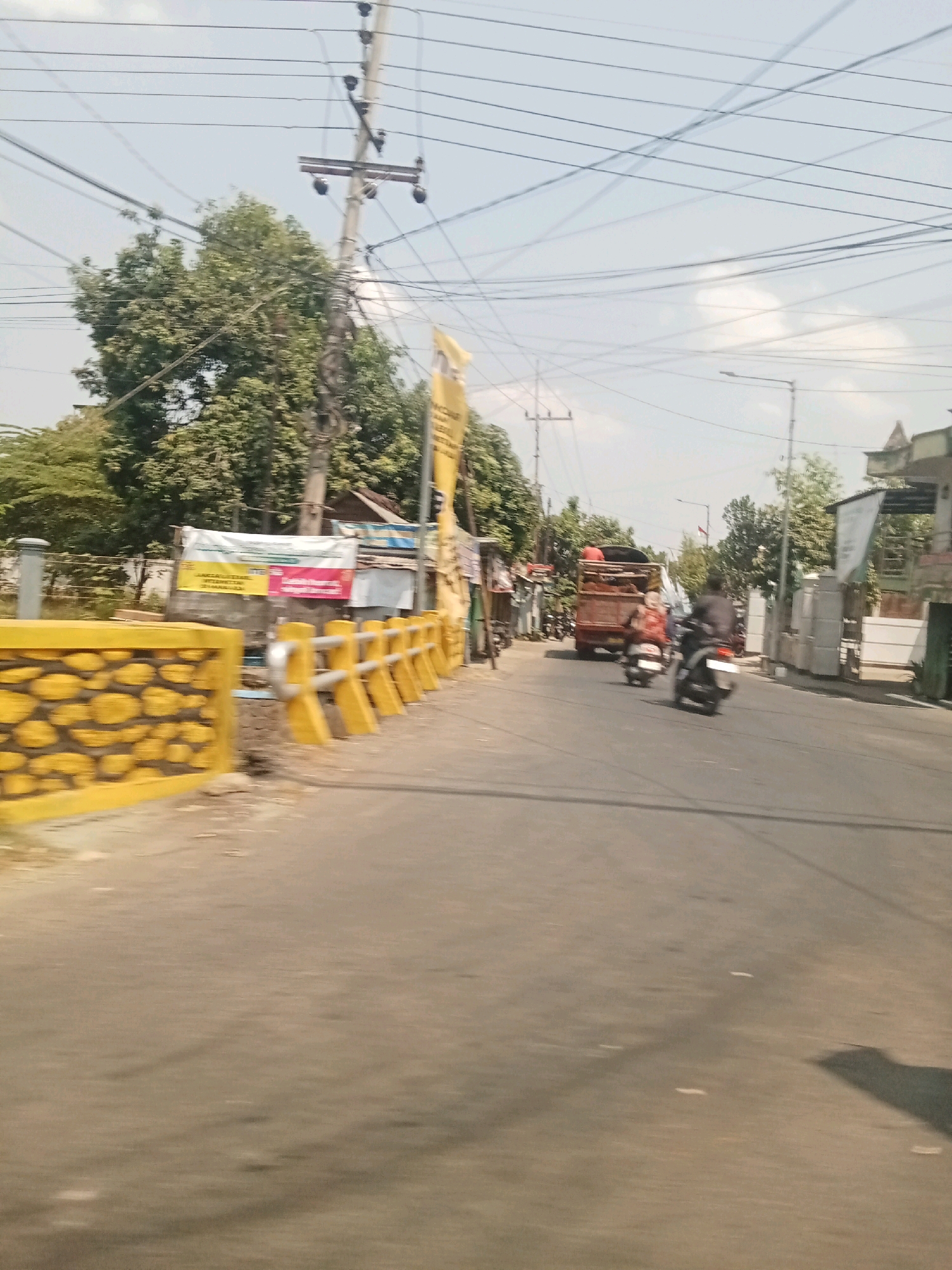 The image depicts a street scene in Tambak Rejo, Indonesia, under a bright, partly cloudy sky. A two-lane asphalt road occupies the foreground, with a yellow guardrail with stone-like texture on the left and a yellow metal guardrail with reflective bars on the right. Several power lines crisscross the sky, supported by utility poles.
On the left side of the road, there are trees, a building with a white banner, and what appears to be a small shop or stall. A tall yellow banner with Indonesian text is also visible. On the right side of the road, there are more trees, buildings, and a street lamp.
The road shows signs of traffic. Two motorcycles are moving away from the viewer, with one rider and a passenger on the first and a single rider on the second. Further down the road, a large, orange truck with a red bed is visible, also moving away. The overall atmosphere suggests a typical day in a semi-urban or rural area with ongoing activity.