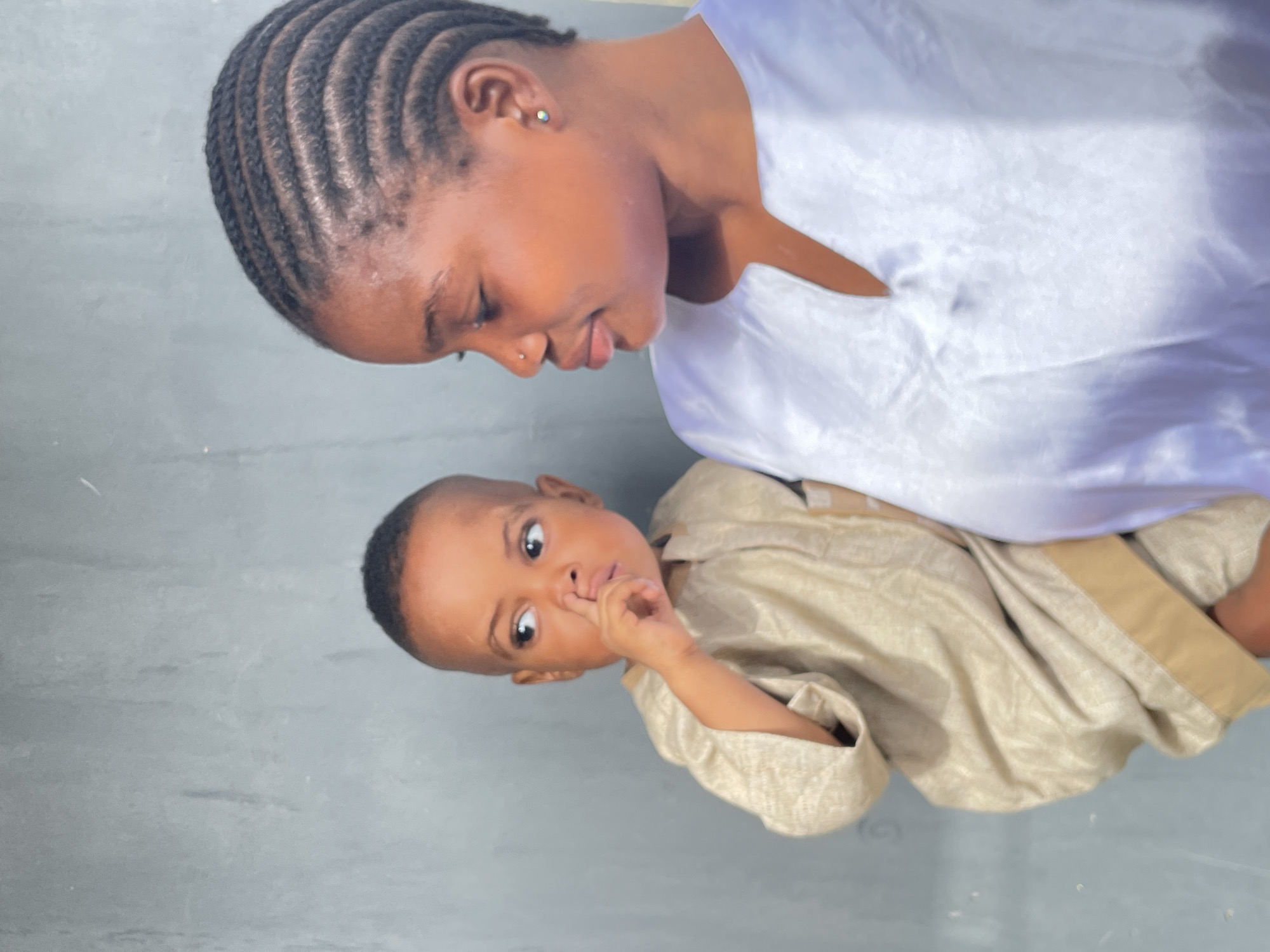 This image captures a tender moment between an adult female and a young child, likely a mother and child, in what appears to be Badagry, Nigeria.
The adult female, positioned on the left side of the frame (when viewed with the subjects upright), is looking down towards the child with a gentle, affectionate expression. She has neatly cornrowed or braided dark hair, a small silver nose piercing, and a silver stud in her ear. She is wearing a simple, light-colored garment, likely white, with a V-neck opening.
The young child, positioned on the right side of the frame, looks directly at the viewer with wide, curious eyes. The child has short, dark hair and is dressed in a light gold or beige traditional-style garment, possibly made of a textured fabric. The child's right hand is raised, with a finger touching their nose or mouth, conveying a pensive, shy, or perhaps questioning gesture.
The background is a plain, light grey surface, possibly a concrete wall or floor. The lighting is bright and natural, indicating that the photo was taken during the daytime. The overall atmosphere of the image is warm and intimate, highlighting the connection between the two individuals. There is no visible text in the image.
