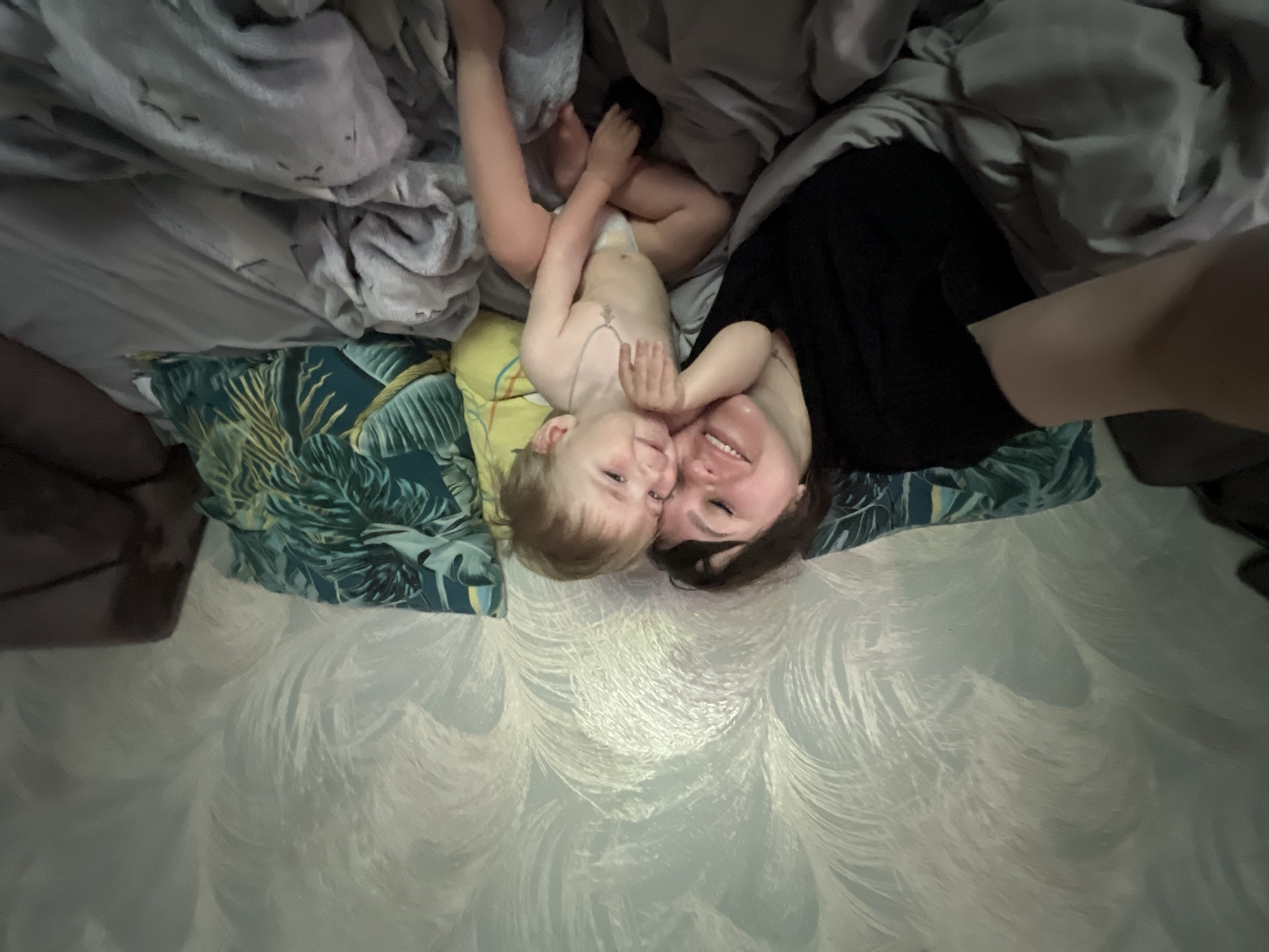 A woman and a young child are in bed. The woman is wearing a black shirt. The child is partially unclothed. They are lying on a bed covered with gray and blue patterned bedding and cushions. The headboard or wall behind the bed is a light blue or white color with a textured or patterned surface. The image appears to be taken from an overhead perspective. The location is not clear enough to identify the city of Noviy, Russia.