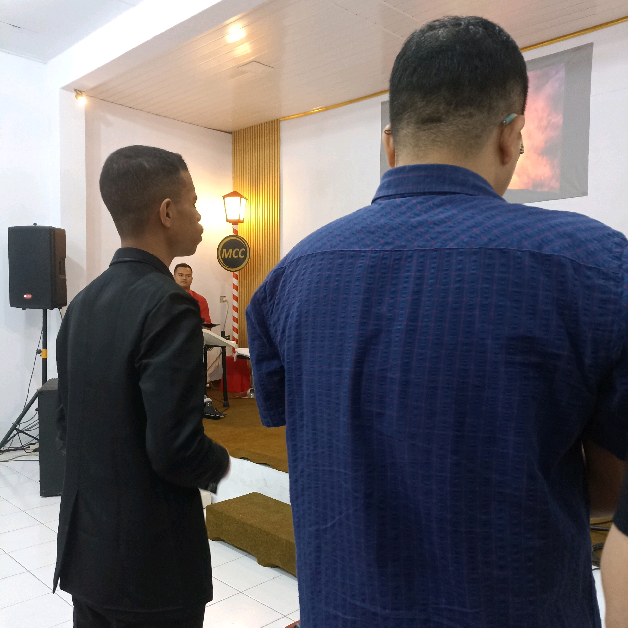 Two men are standing in what appears to be an indoor event space, possibly a church hall or community center, in Magelang, Indonesia. The man on the left, seen from the back, is wearing a black suit. The man on the right, also seen from the back, is wearing a blue patterned shirt. Behind them, a person in a red shirt is seated at a keyboard. A speaker is visible to the left of the frame. In the background, a screen displays an image of fire. A decorative lamp post with a circular sign bearing the letters "MCC" is also present. The floor is tiled, and the walls and ceiling are white. The overall atmosphere suggests a gathering or performance.