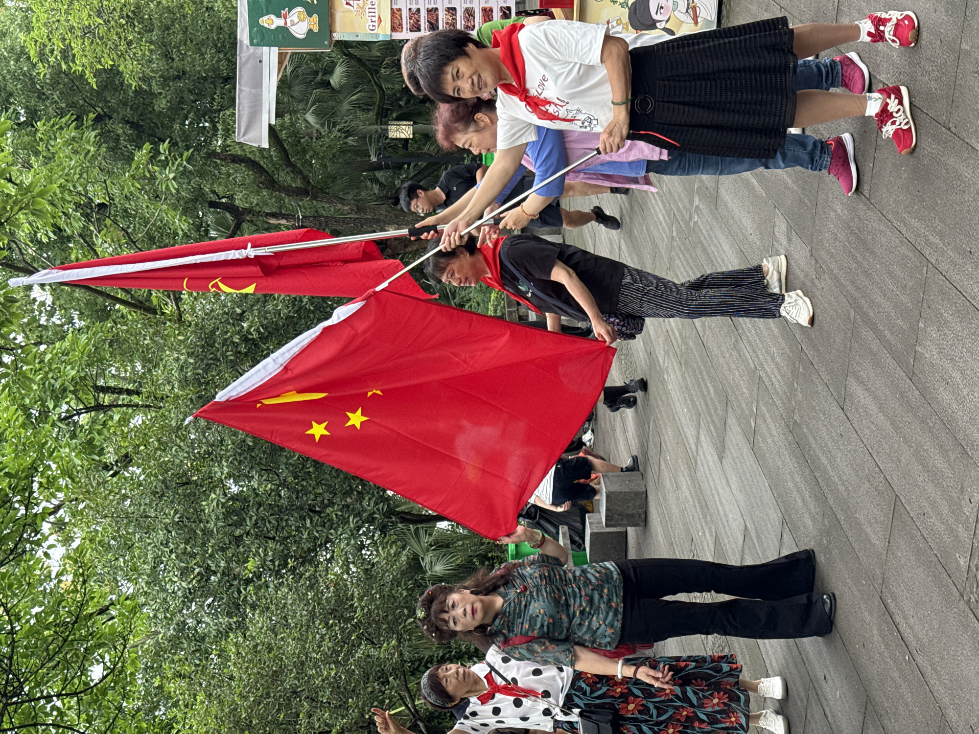 A group of approximately 10 adults are on a paved walkway, holding two Chinese flags. The people are of various ages, and are wearing casual clothing. They appear to be in a park or public area in Yuzhong District, China. A menu for a restaurant is visible in the background. The walkway is gray and tiled. There are trees behind the group.