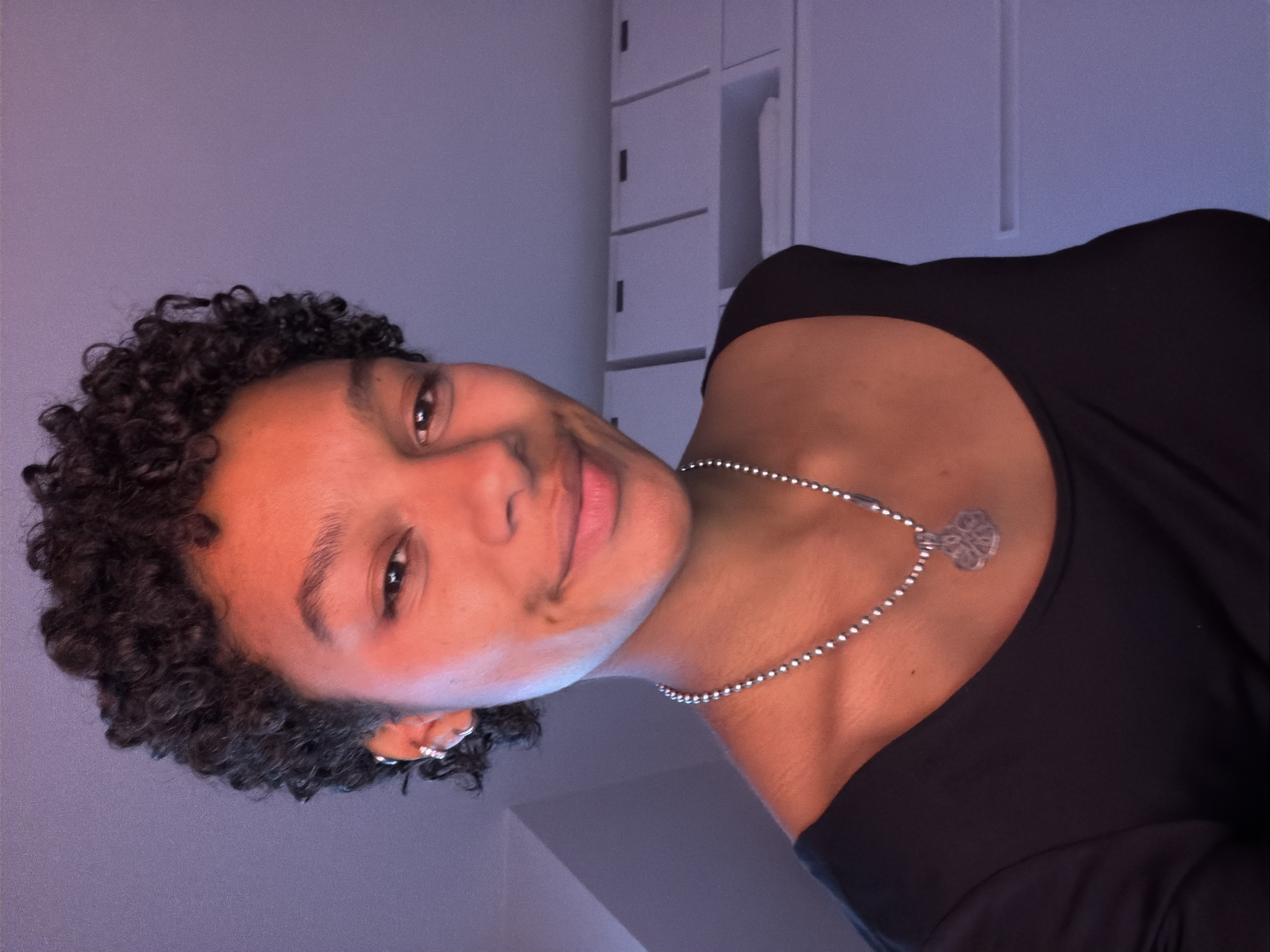 A medium-shot, rotated portrait shows a woman with curly, dark hair, light brown skin, and a slight smile. She wears a black top and a silver necklace with a pendant. She has a ring on her left ear. In the background, there is a white storage cabinet and a purple wall. There is no information provided to determine the location of this image within Paris, France.