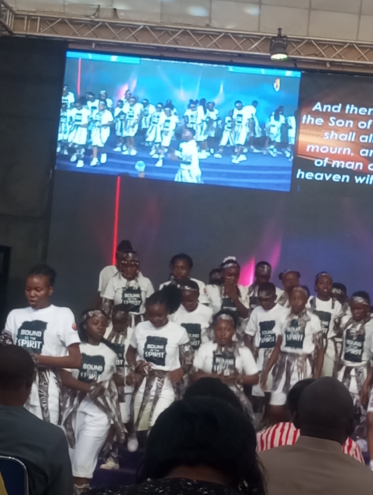 A group of children and adults wearing white T-shirts with "SOUND OF THE SPIRIT" printed on them stand on a stage in what appears to be a church or auditorium. They also wear striped silver and white wraps around their waists and on their heads. Behind them, a large screen shows another group of people, similarly dressed, performing. Part of a biblical verse is visible on the right side of the screen. People are seated in the foreground, facing the stage. The location appears to be Gura Top, Nigeria.