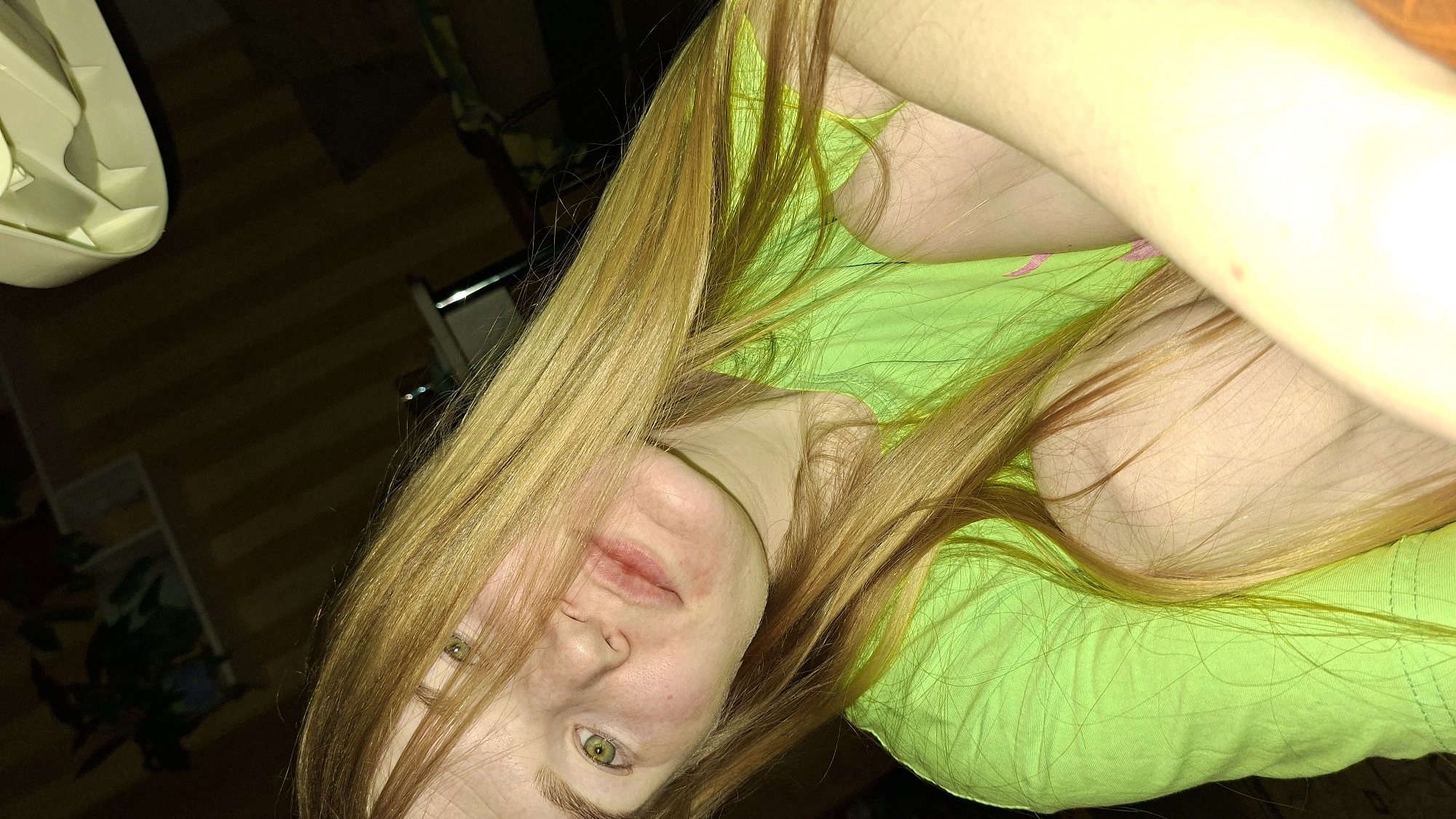 A close-up, low-angle shot depicts a person with long, light golden-brown hair. The person is wearing a bright green garment. The lower portion of the face shows visible redness around the mouth and nose. The eyes are a light green color. Background details include a section of a wooden floor and objects that appear to be furniture or storage containers. The location is unknown.
